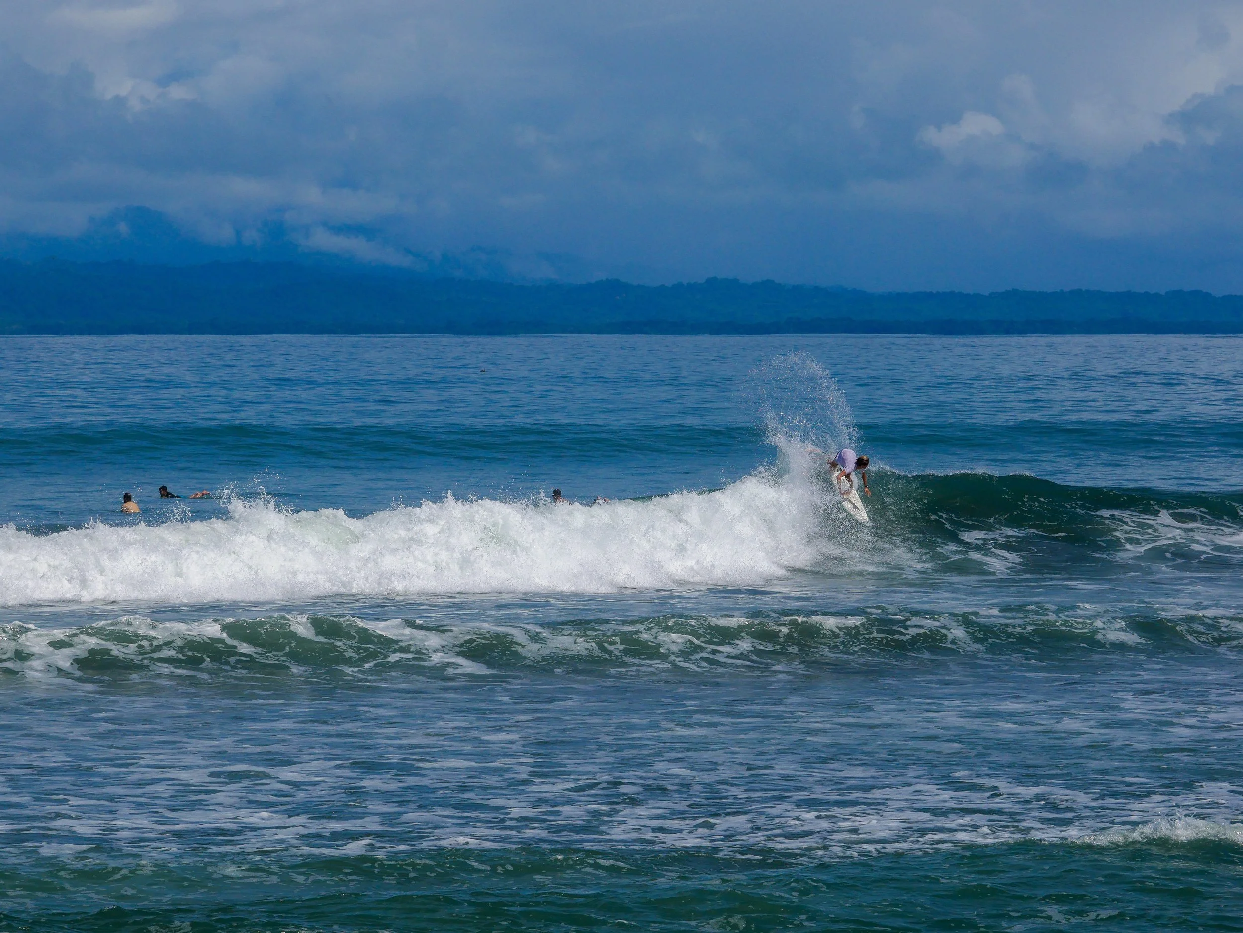 Surfer riding a wave in the ocean with other people swimming nearby under a cloudy sky.