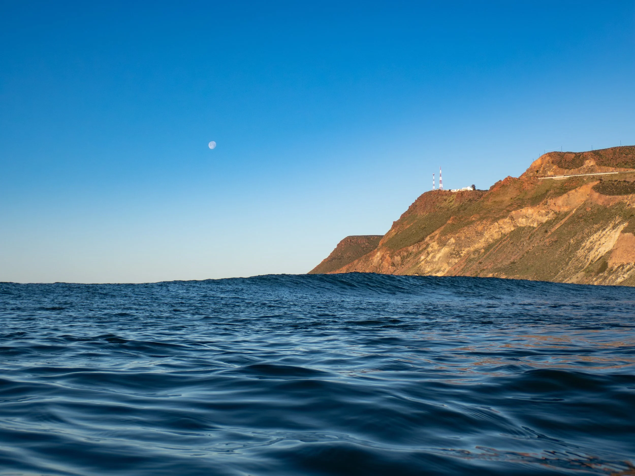 Ocean water with a distant view of a rocky hill or mountain with antennas and a building on top, under a clear blue sky with visible moon.