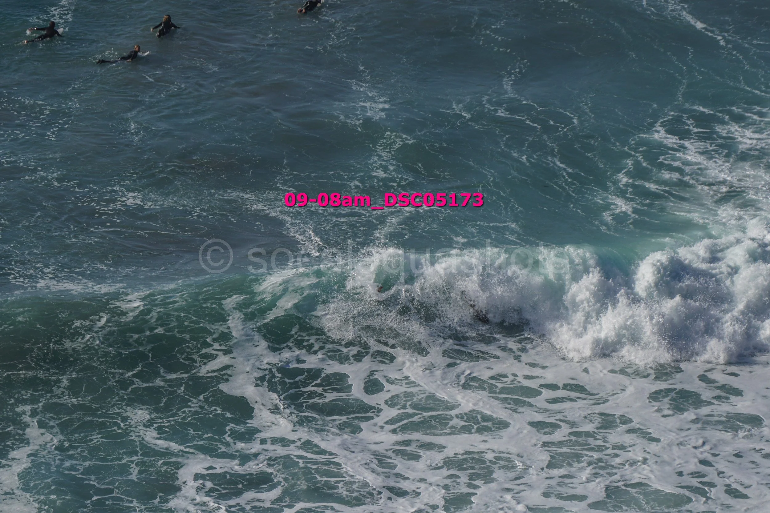 People in wetsuits swimming in the ocean near a large breaking wave.