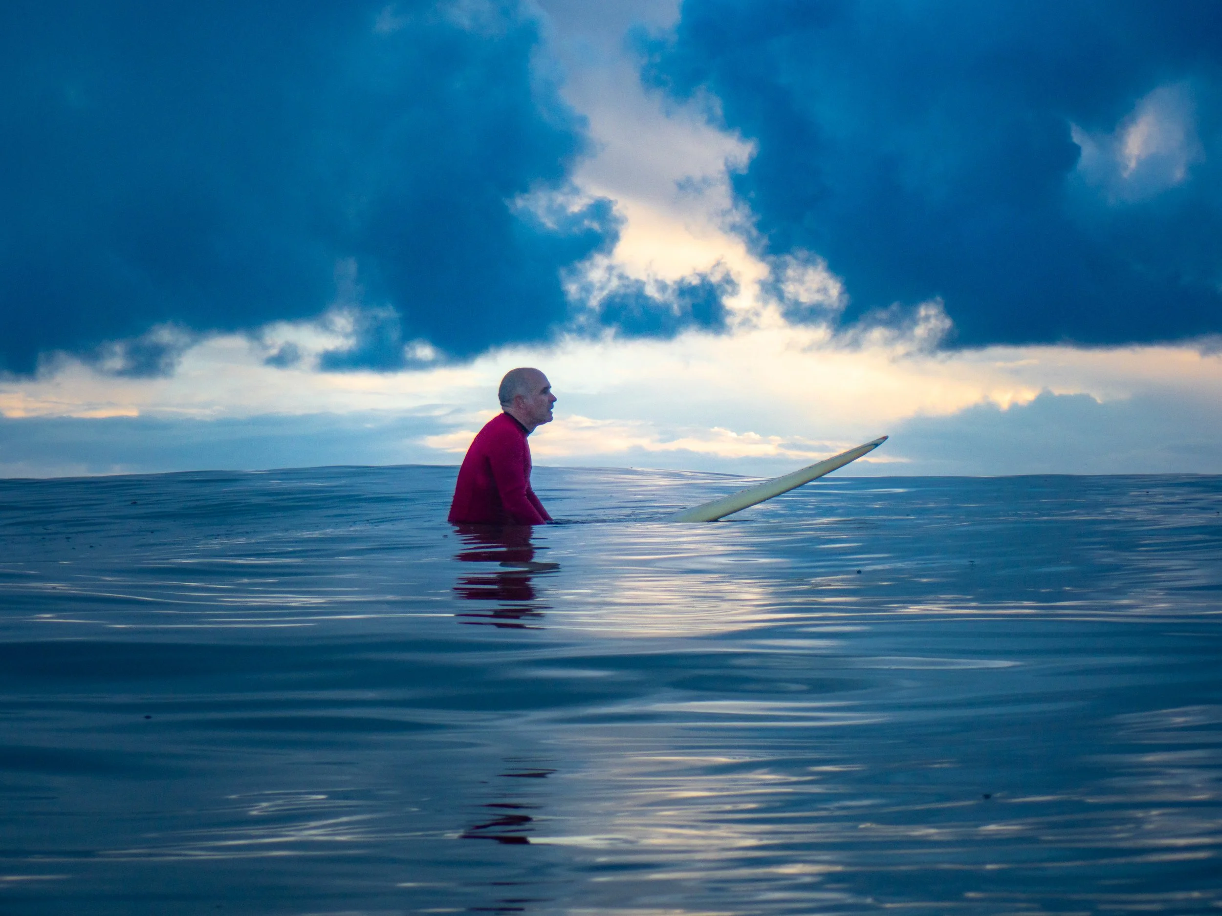 An elderly man with gray hair in a red sweater standing waist-deep in calm water, looking at a surfboard floating nearby under a cloudy sky during sunset.