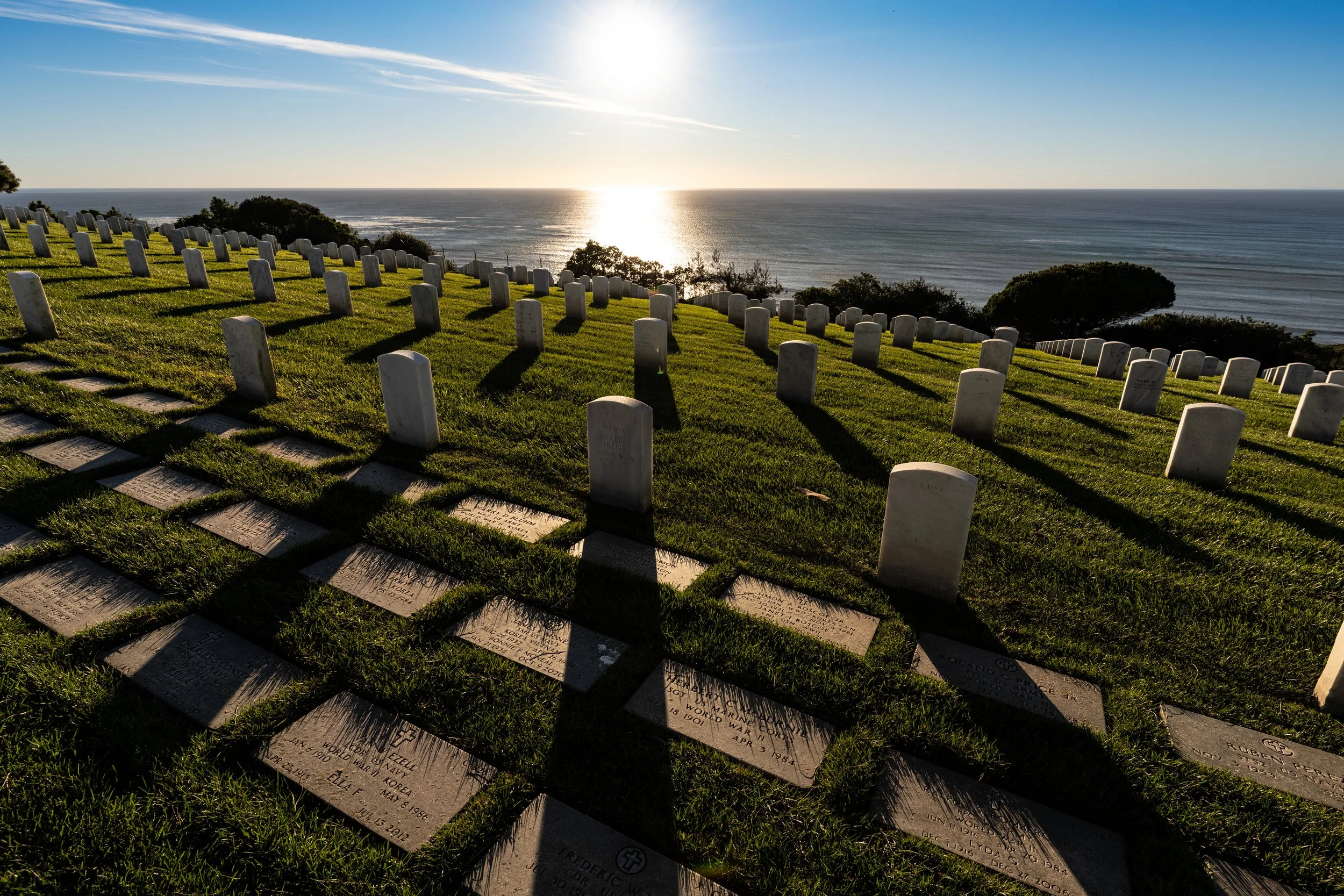 A cemetery on a hillside overlooking the ocean during sunset, with numerous white headstones casting long shadows on green grass.