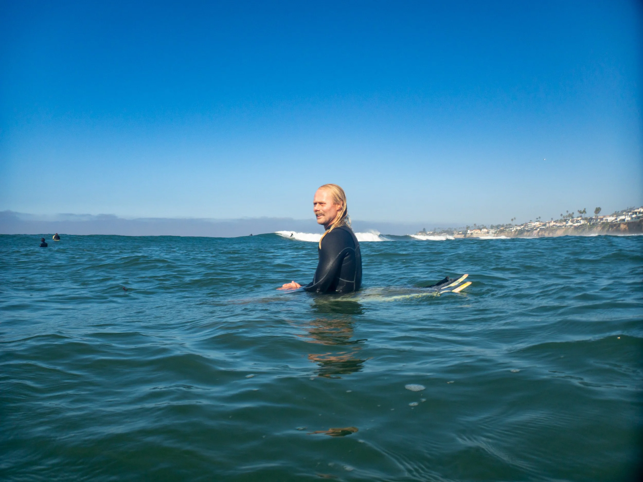 Man in wetsuit sitting on surfboard in the ocean, looking at the camera, with waves and a shoreline with houses and palm trees in the background, under a clear blue sky.
