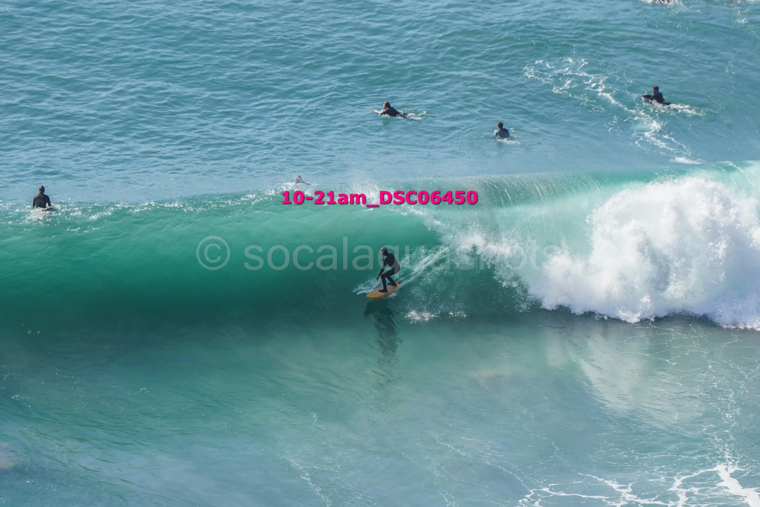Surfer riding a large wave with several people surfing and swimming in the water nearby.