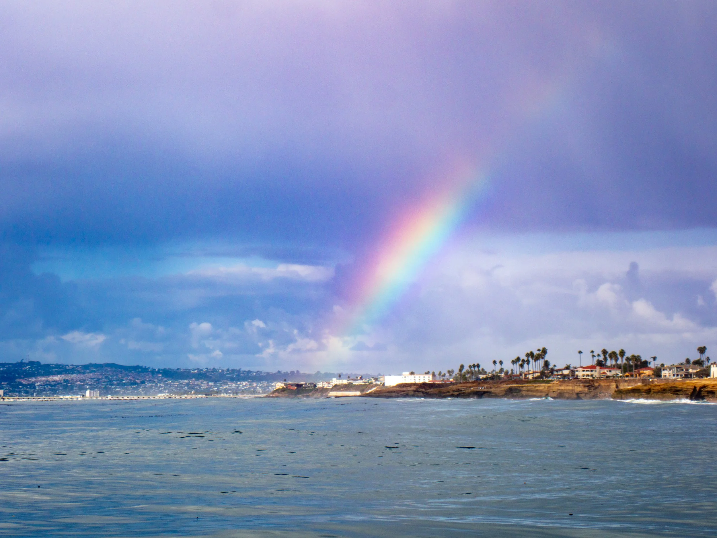 Rainbow over the ocean with a coastal city and palm trees in the background under a cloudy sky.