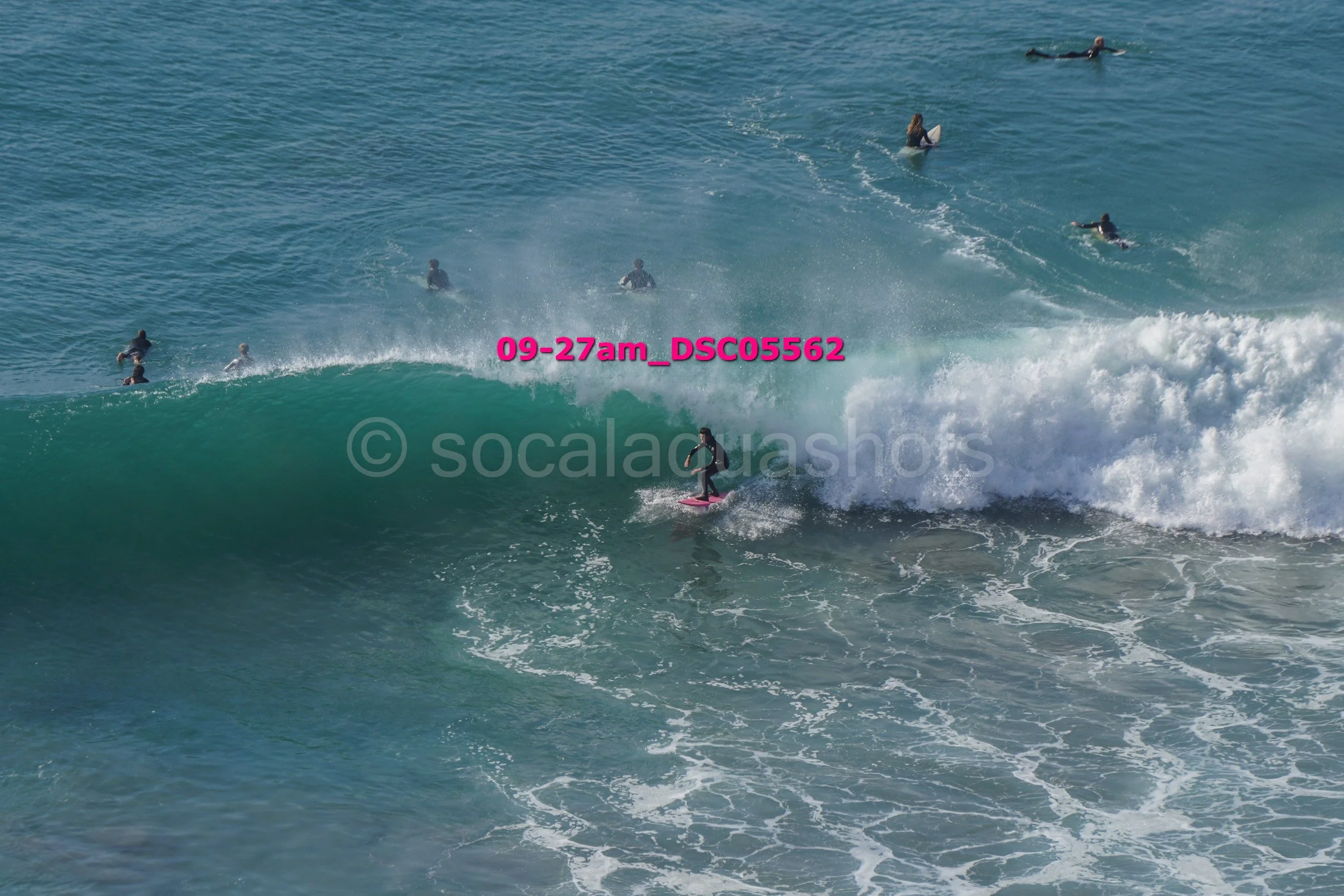 A person surfing on a pink surfboard riding a wave with several observers in the water nearby.