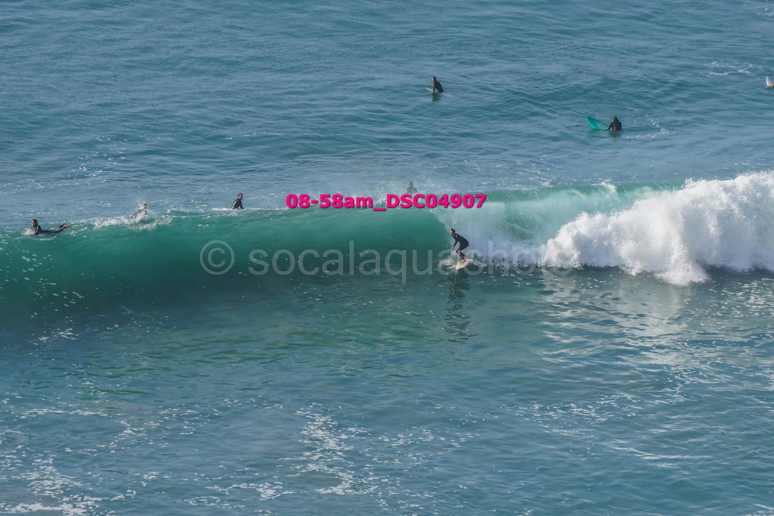 Surfers riding and paddling in the ocean waves, some on surfboards, with a clear blue sky.