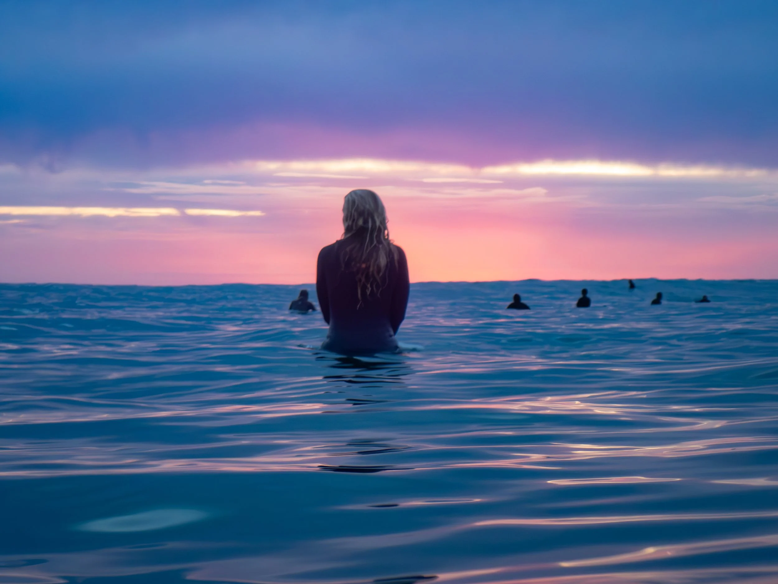A woman with long hair stands in the ocean watching a sunset, with other people visible in the distance.