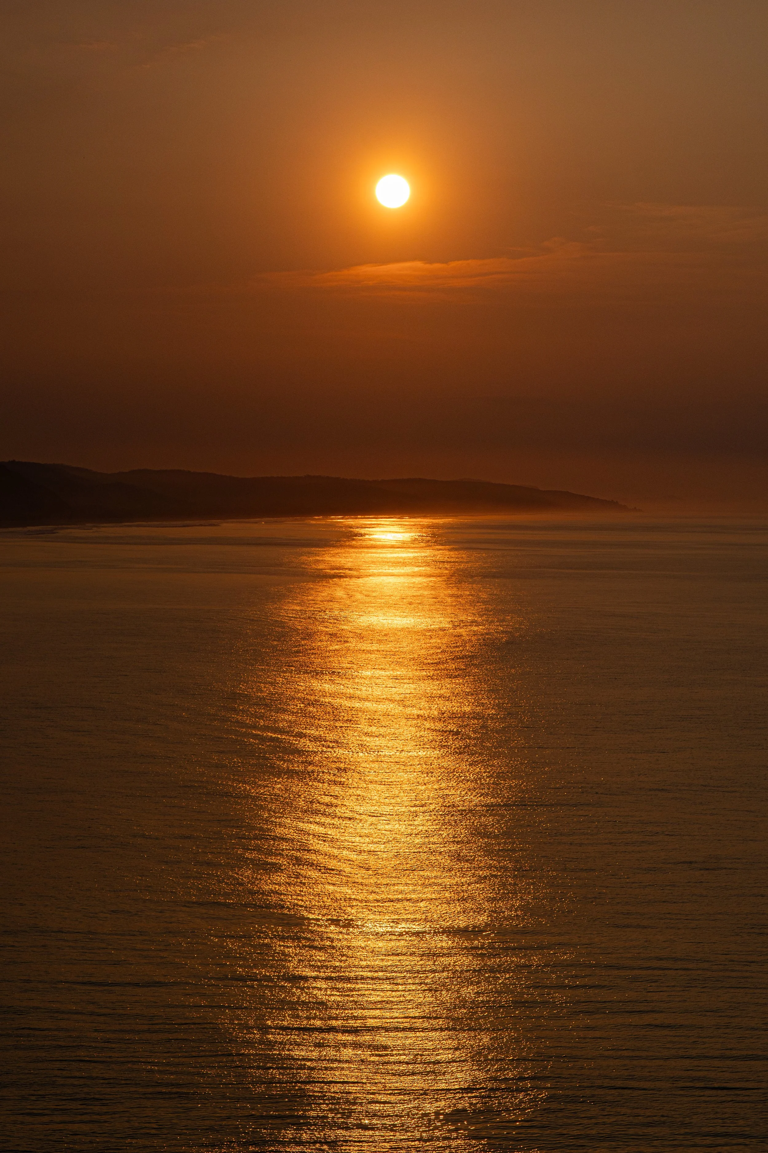 A sunset over the ocean with the sun reflecting on the water and an island or landmass in the distance.