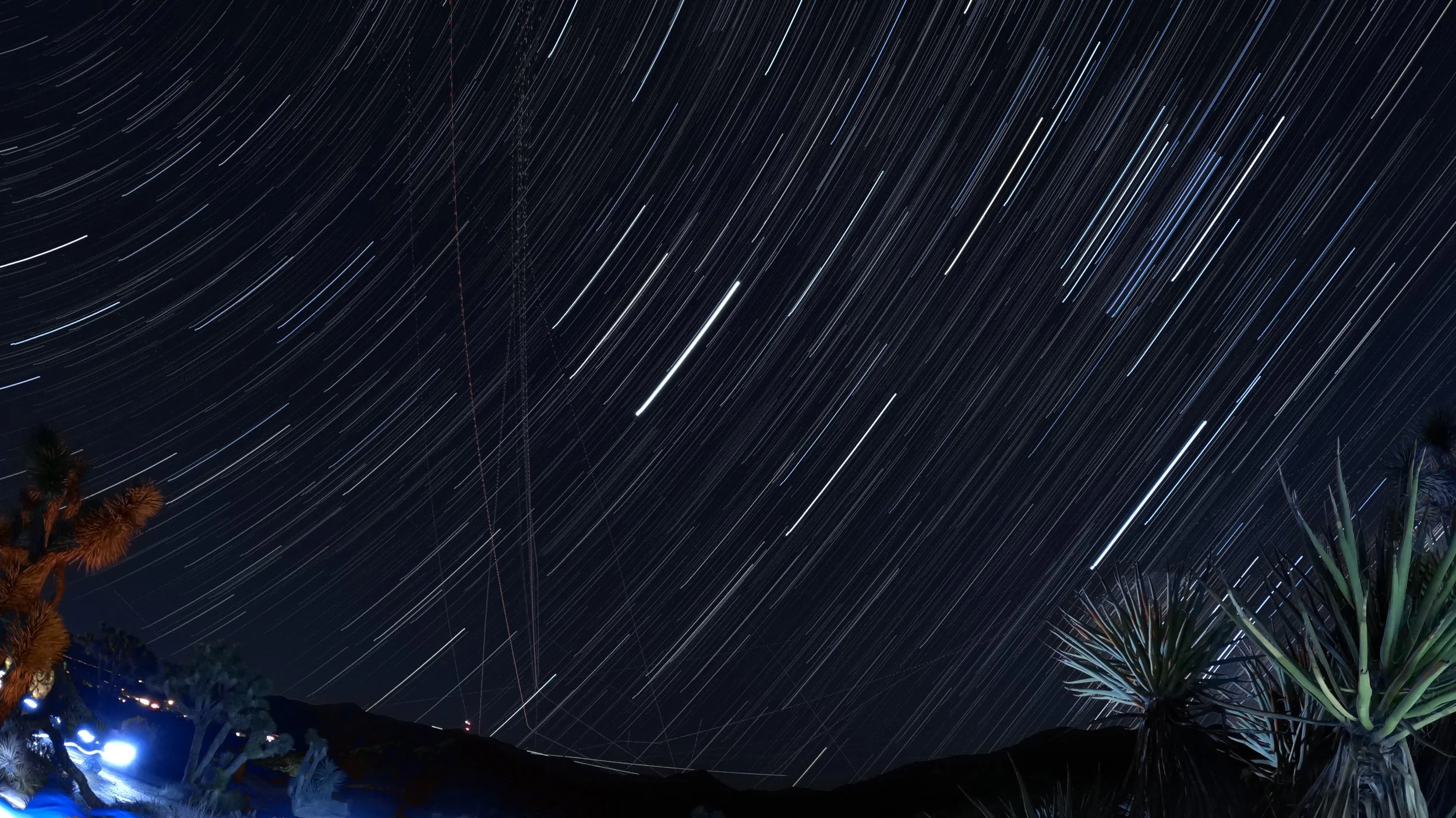 Long exposure photograph of the night sky with star trails and silhouettes of trees and mountains in the foreground.