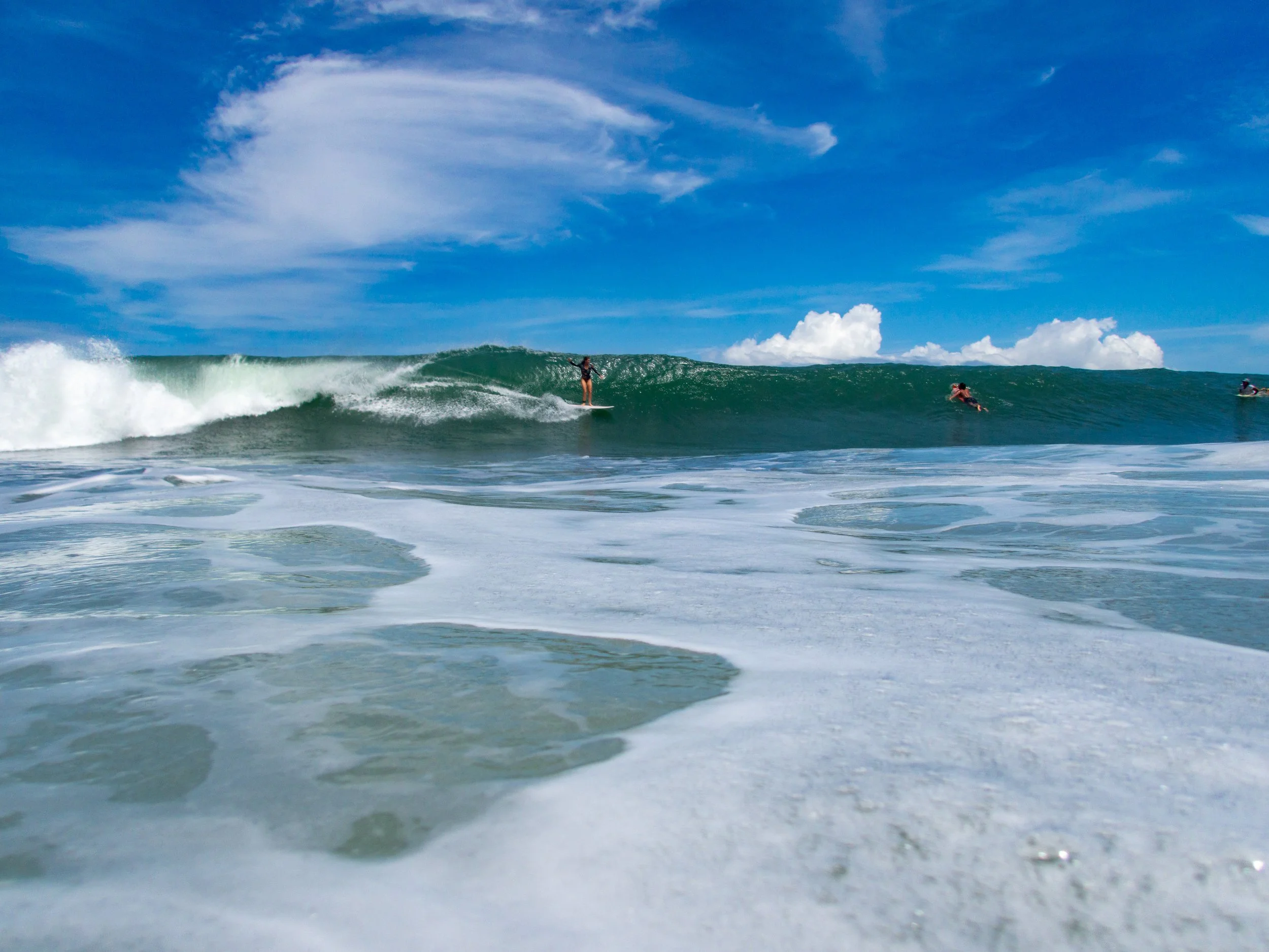 Surfer riding a wave in the ocean under a blue sky with clouds, viewed from water level.