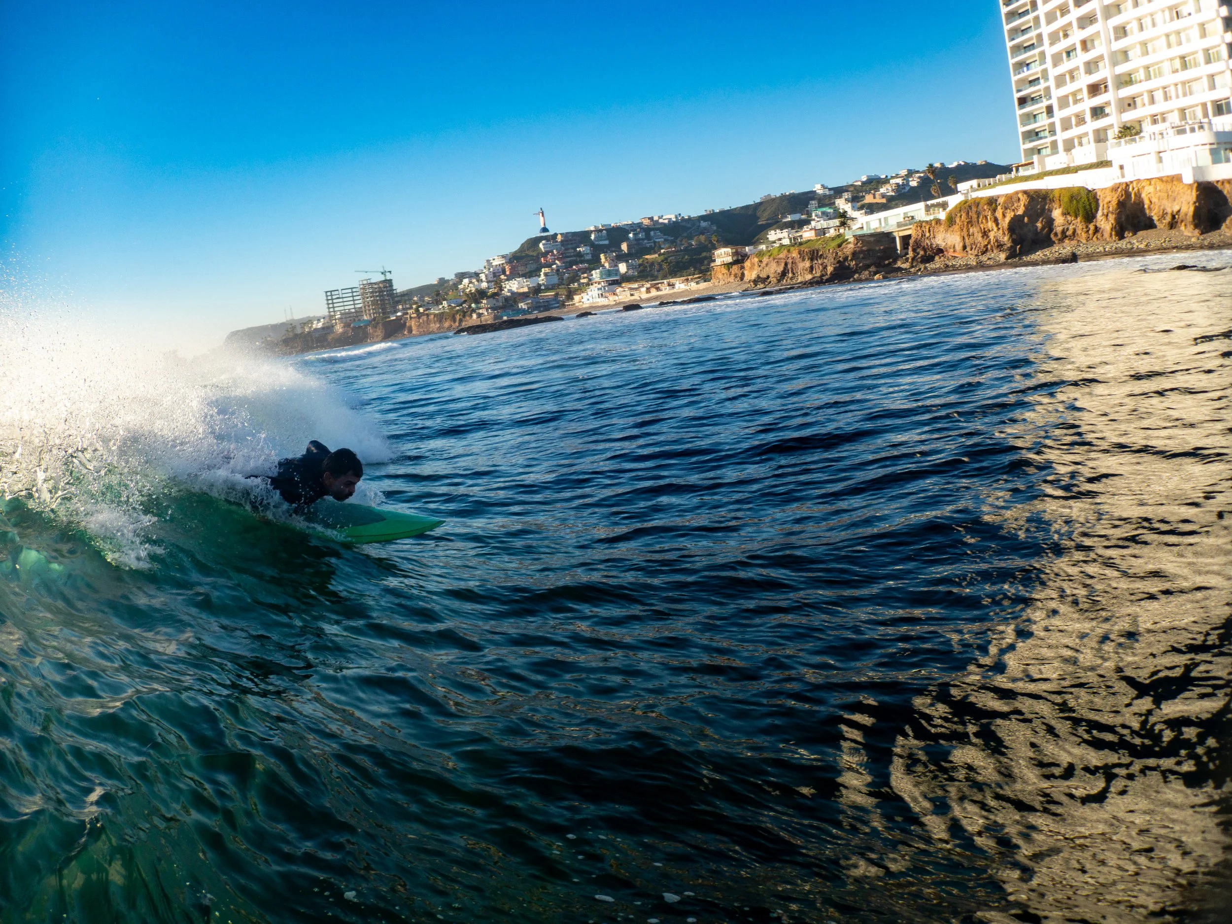 A person surfing on a wave near a coastal shoreline with buildings and cliffs in the background.