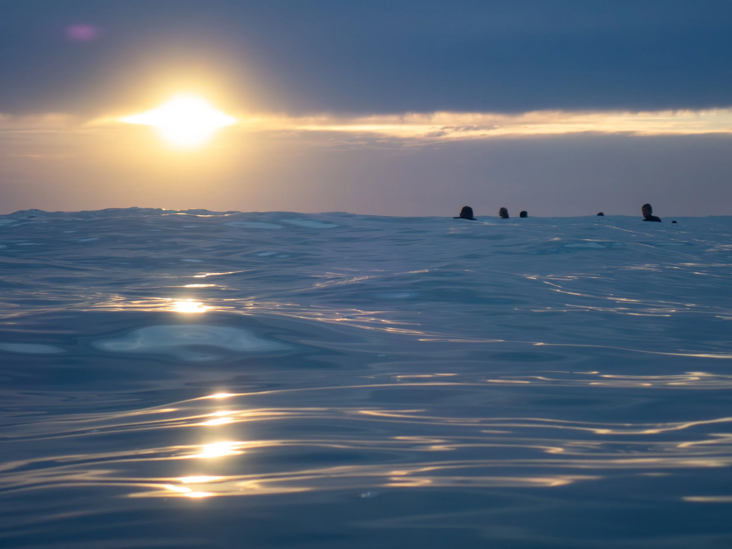 People swimming in the ocean during sunset with a mostly cloudy sky.