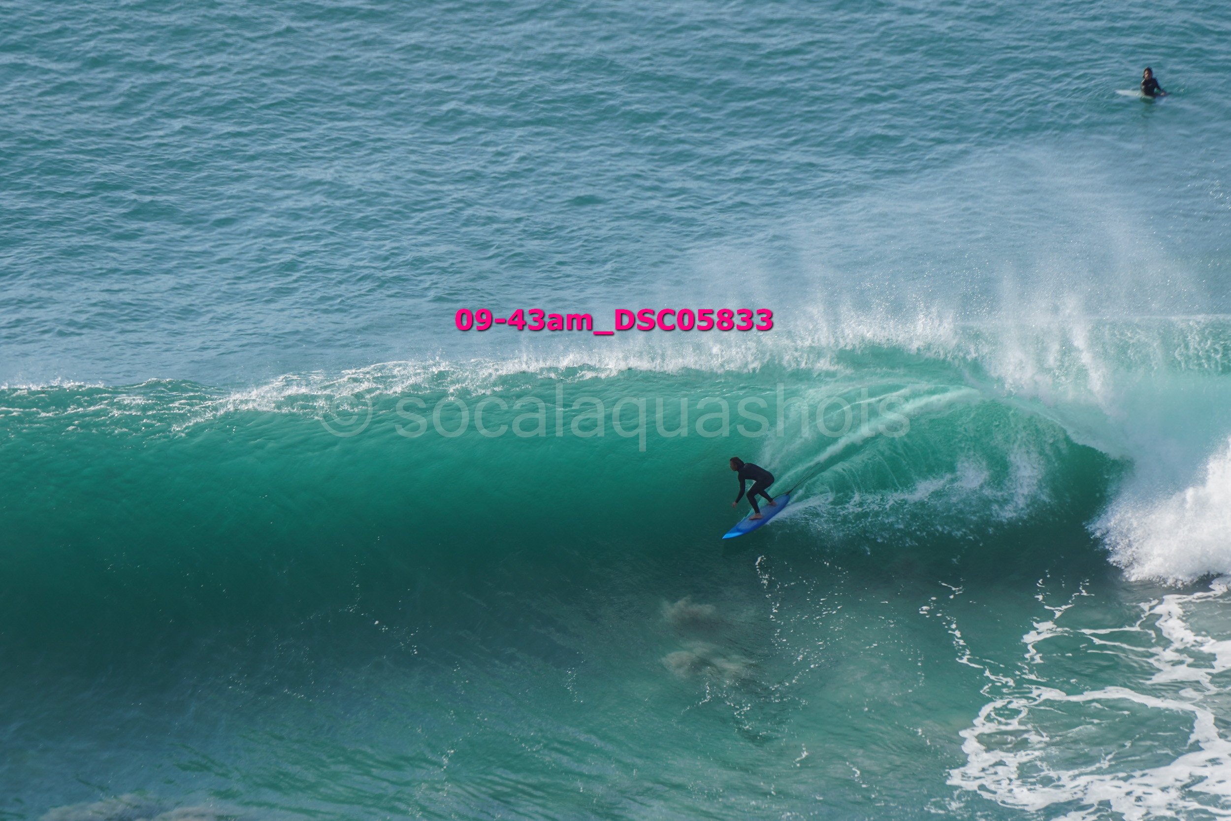 A person surfing on a large ocean wave, with another surfer visible in the distance.