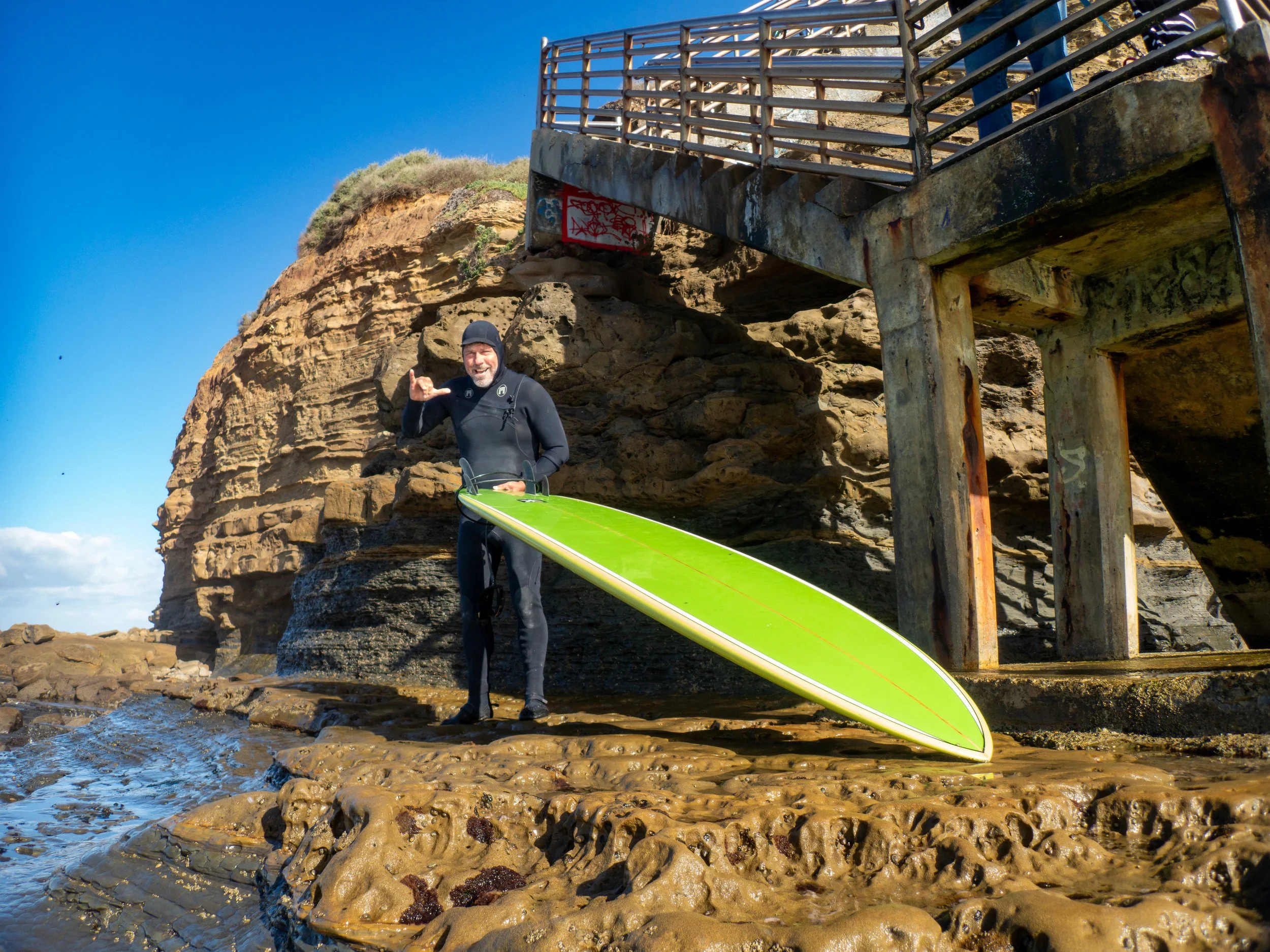 A man in a wetsuit holding a surfboard under a bridge, standing on rocky shoreline with a clear blue sky.