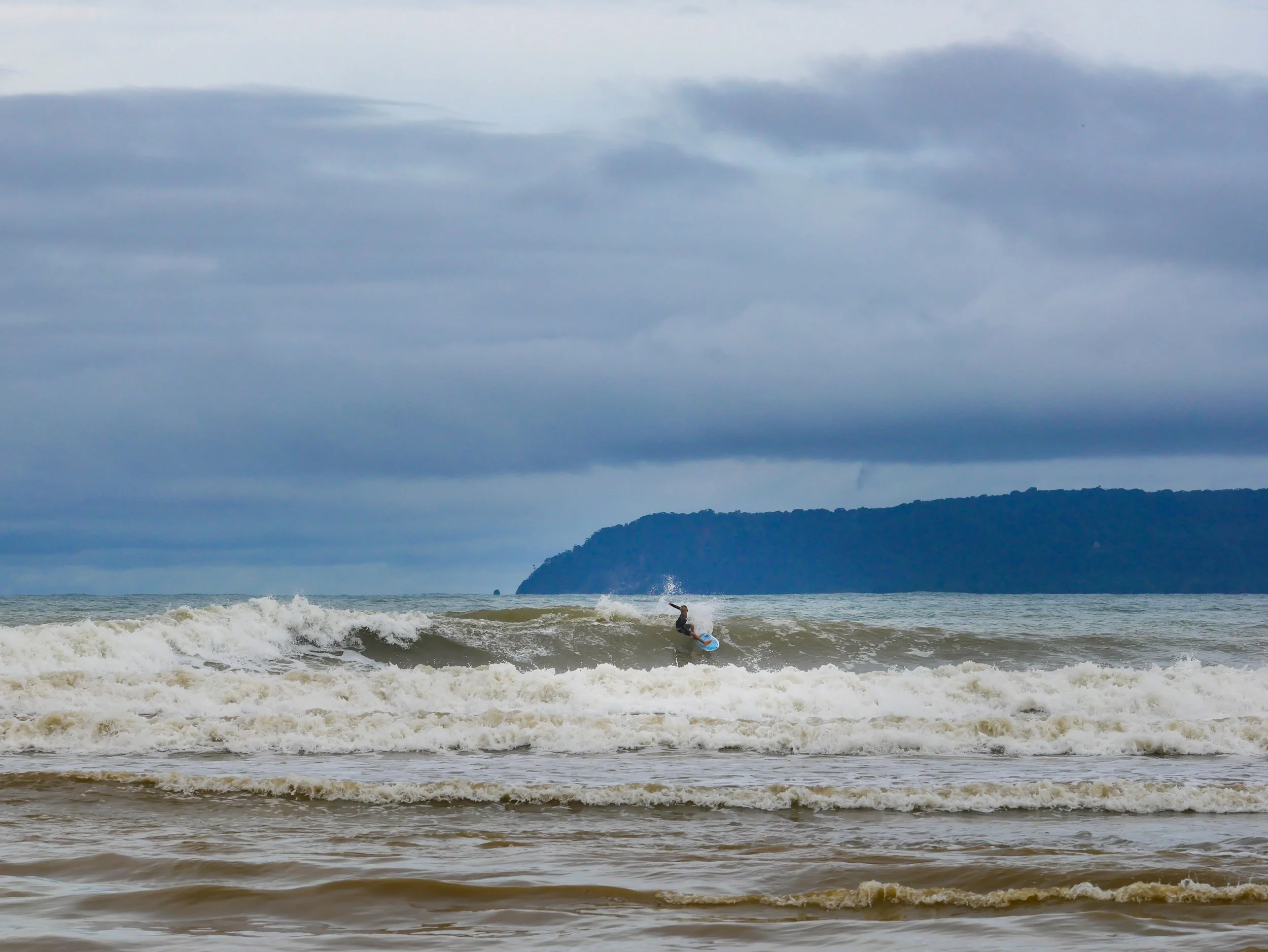 Surfer riding a wave in the ocean with an overcast sky and distant landmass in the background.