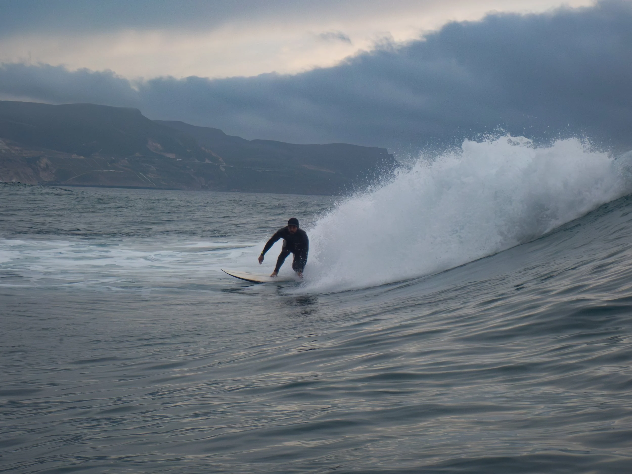 A person surfing on a wave in the ocean with mountains in the background and a cloudy sky.