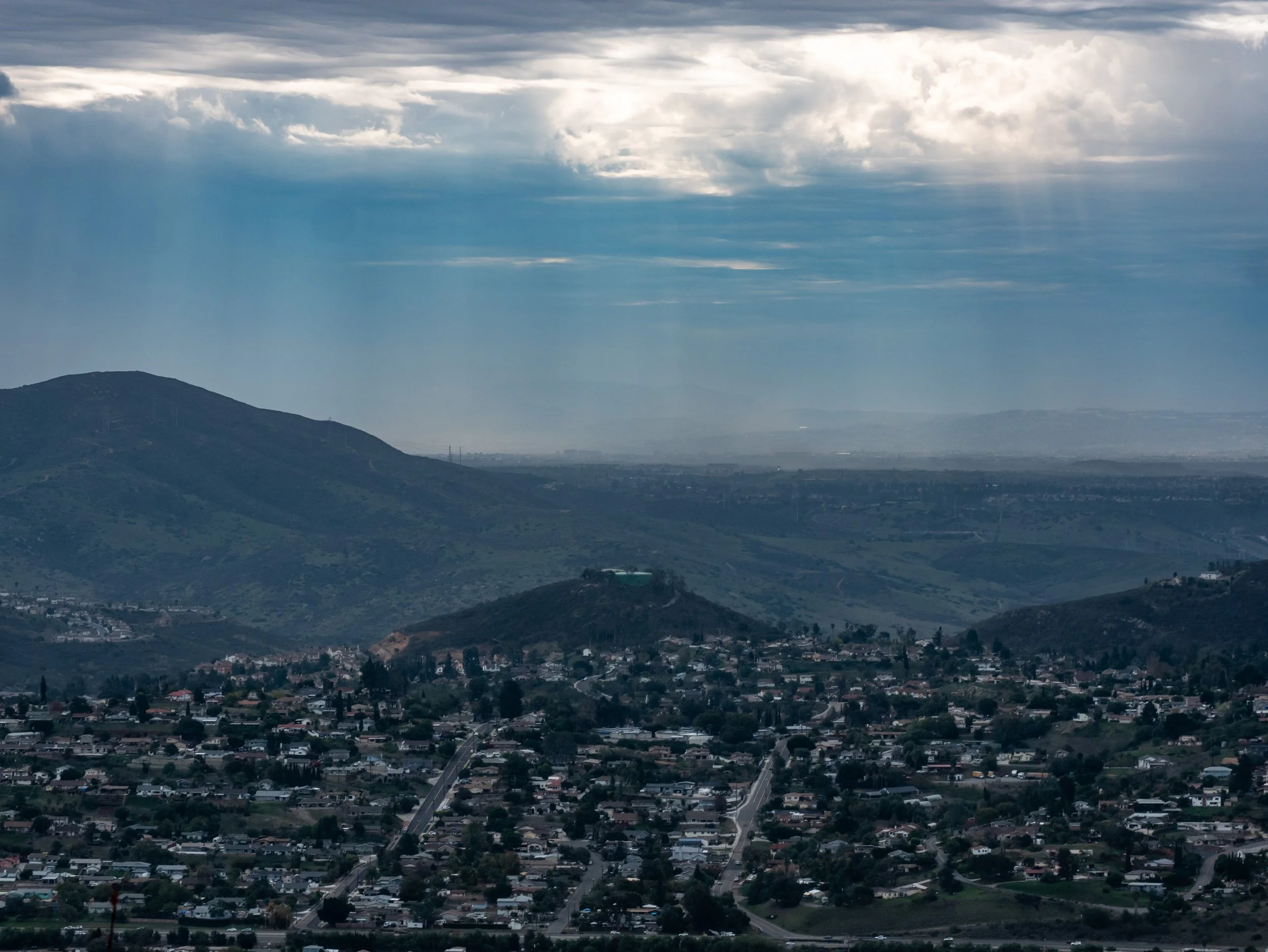 A landscape view of a city with residential houses and streets in the foreground, rolling hills in the middle ground, and mountains in the background. The sky is partly cloudy with rays of sunlight filtering through.