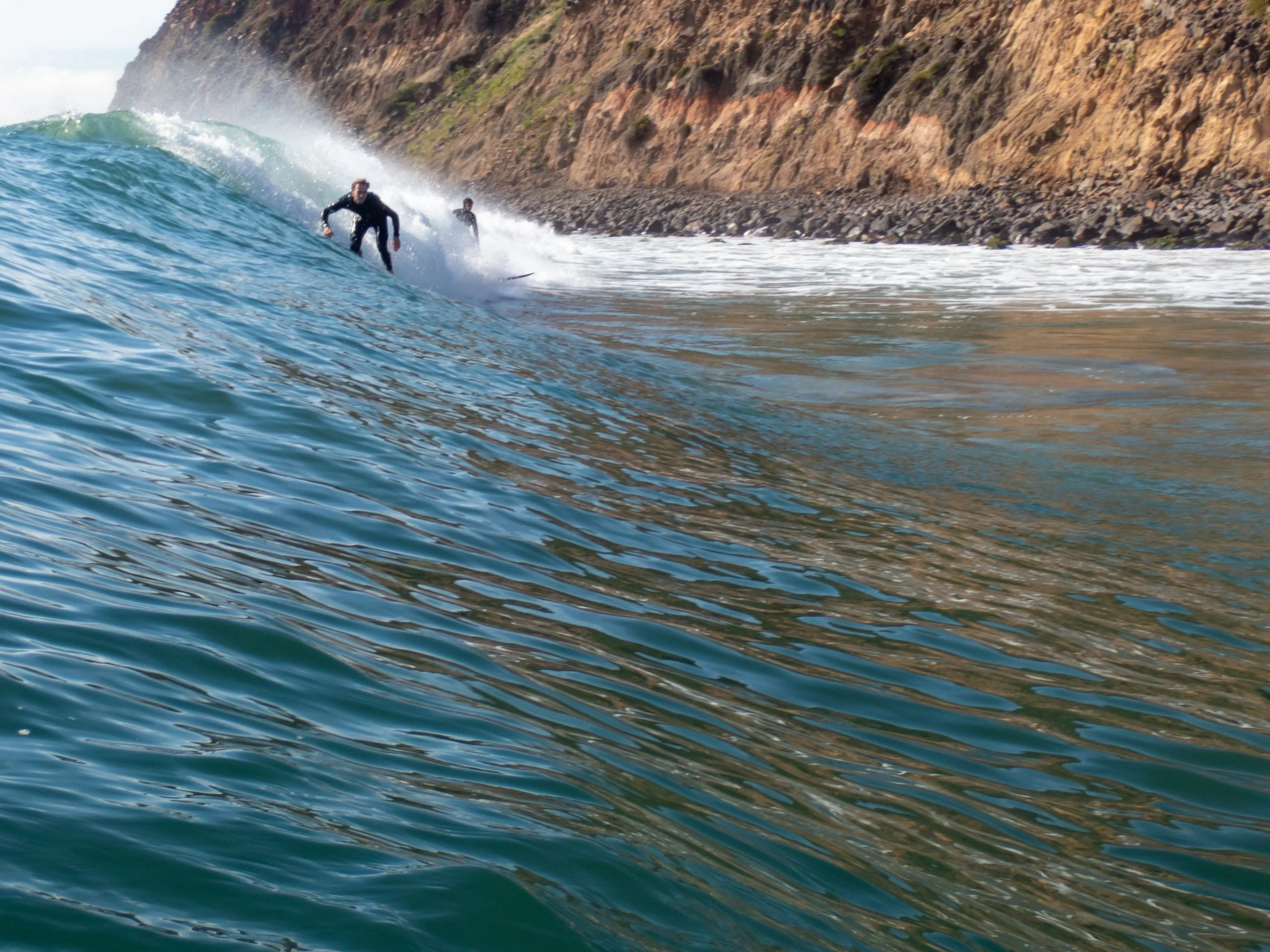 Surfers riding waves near a rocky coastline with cliffs in the background.