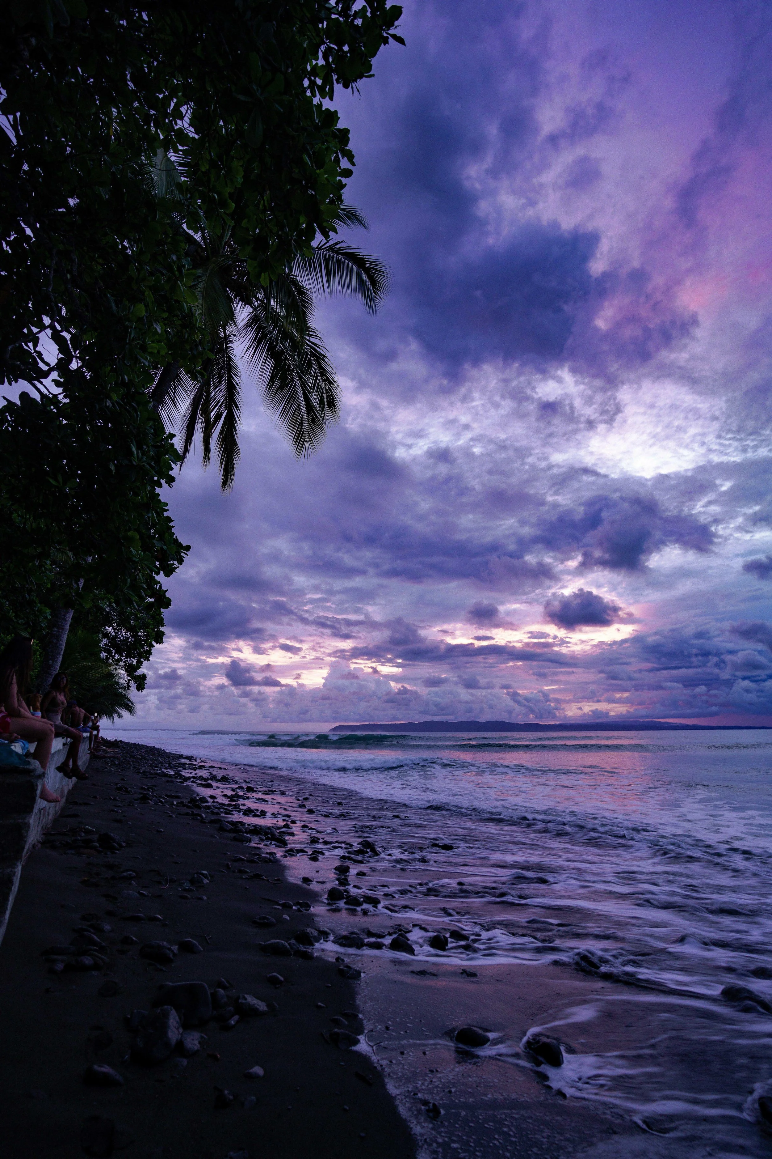 A beach scene at dusk with purple and blue sky, clouds, and calm ocean waves. On the left, there are silhouetted trees and people sitting on a wall, and towards the horizon, a landmass is visible.