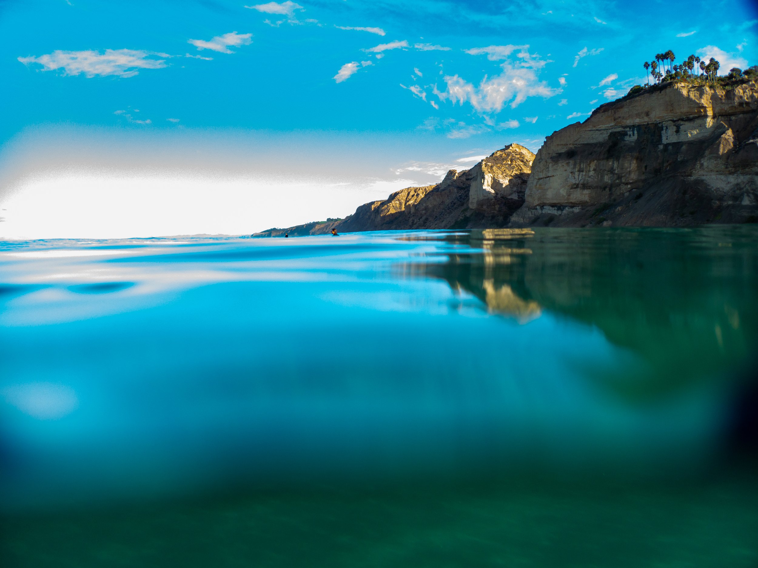 Scenic view of the ocean with clear blue water, rocky cliffs with palm trees on top, and a bright sky with few clouds.