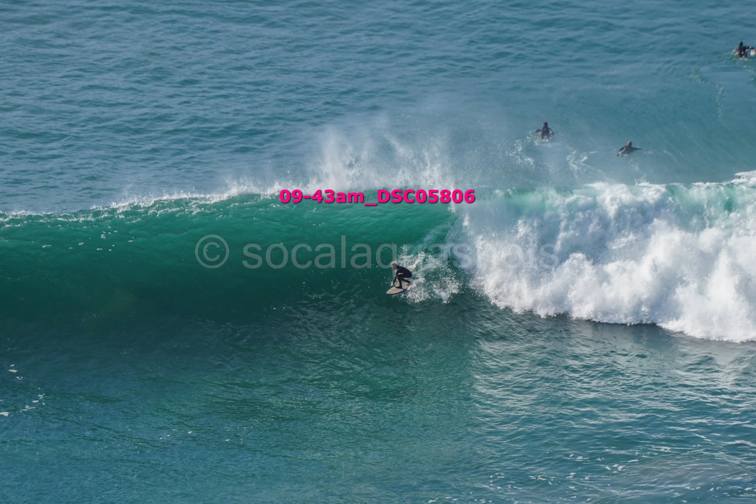 Surfer riding a large wave in the ocean with other surfers visible in the distance.