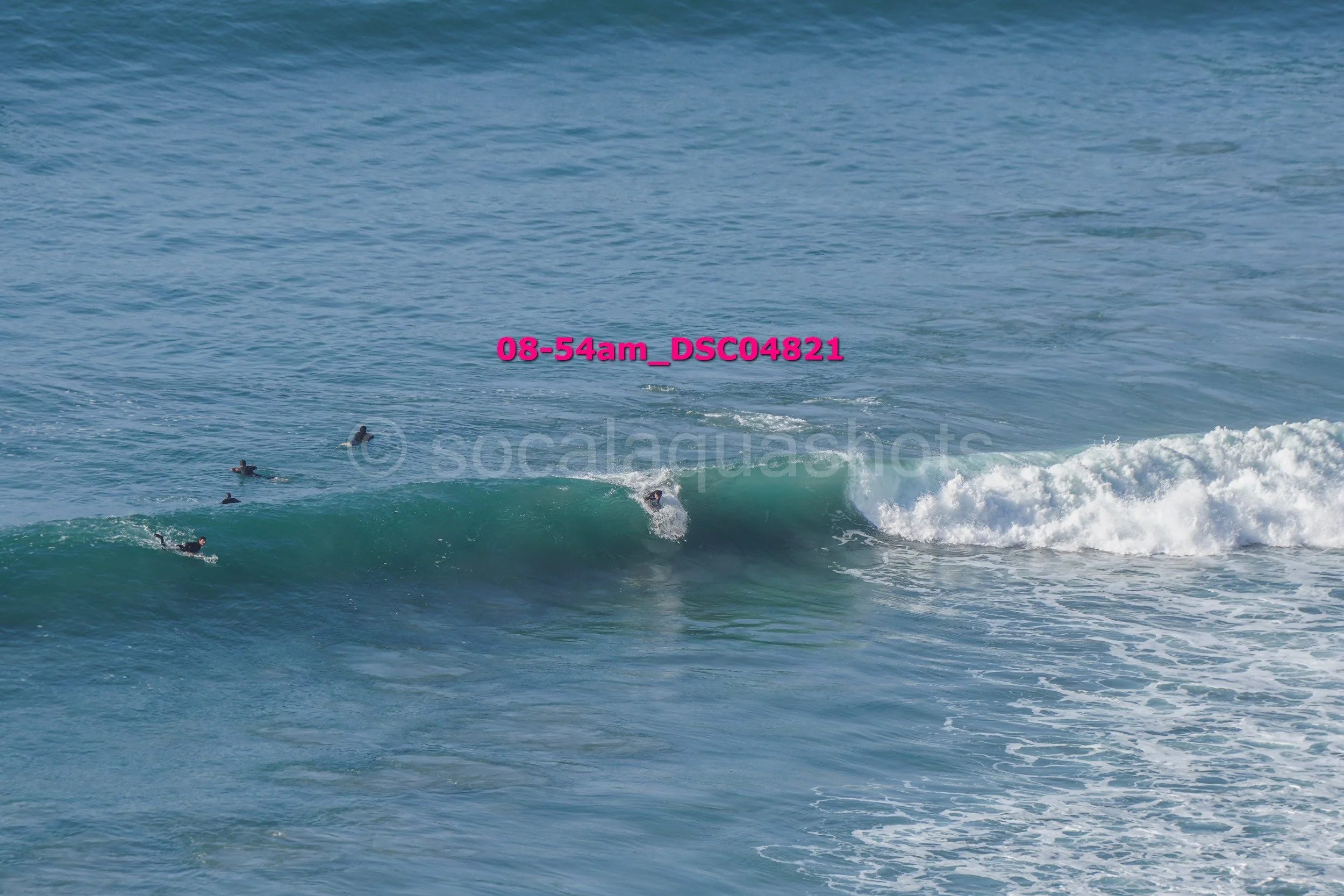 A person surfing on a blue wave with several other surfers swimming nearby in the ocean.