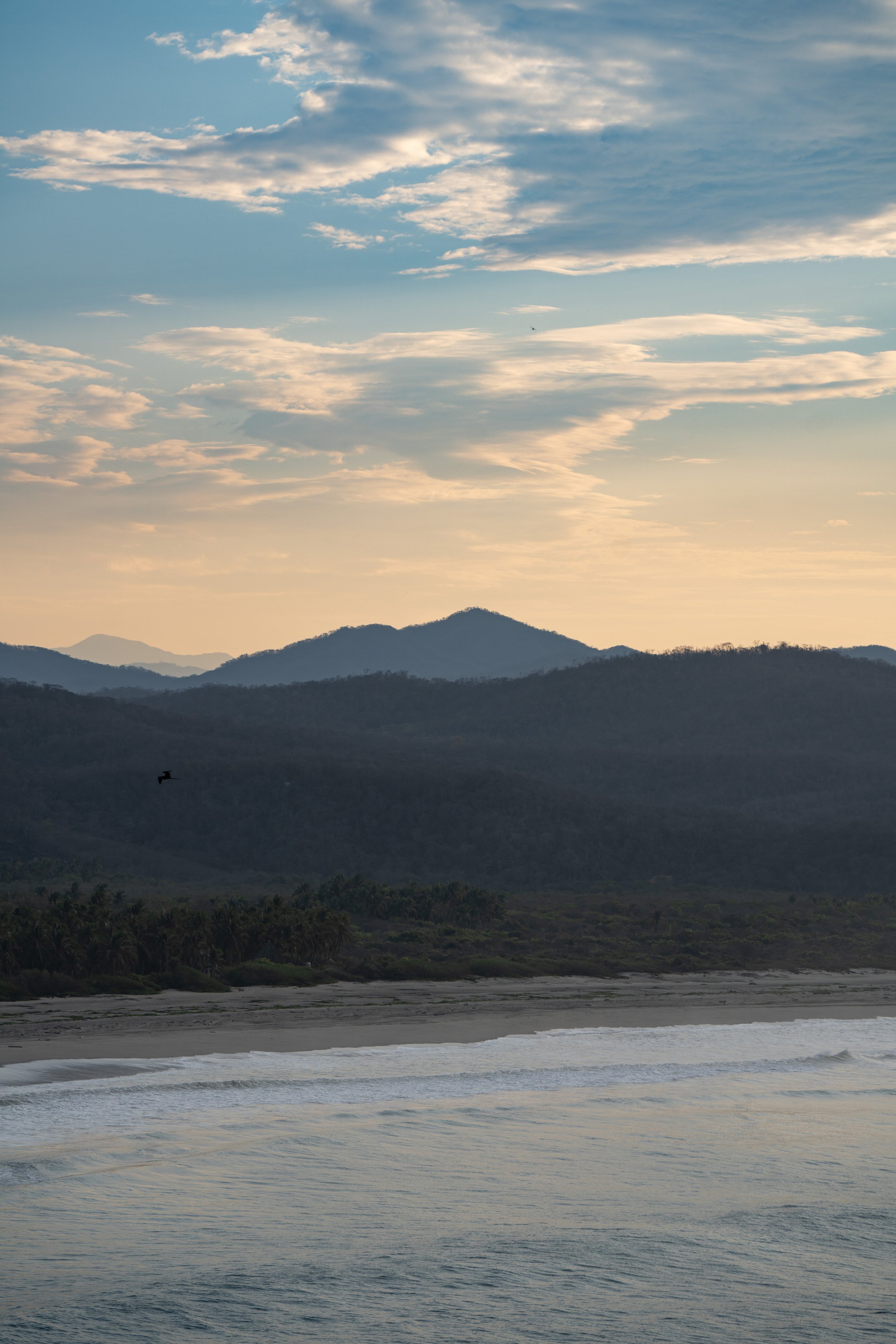 A scenic view of a coastline with calm water in the foreground, rolling hills and mountains in the background, and a partly cloudy sky at sunset.