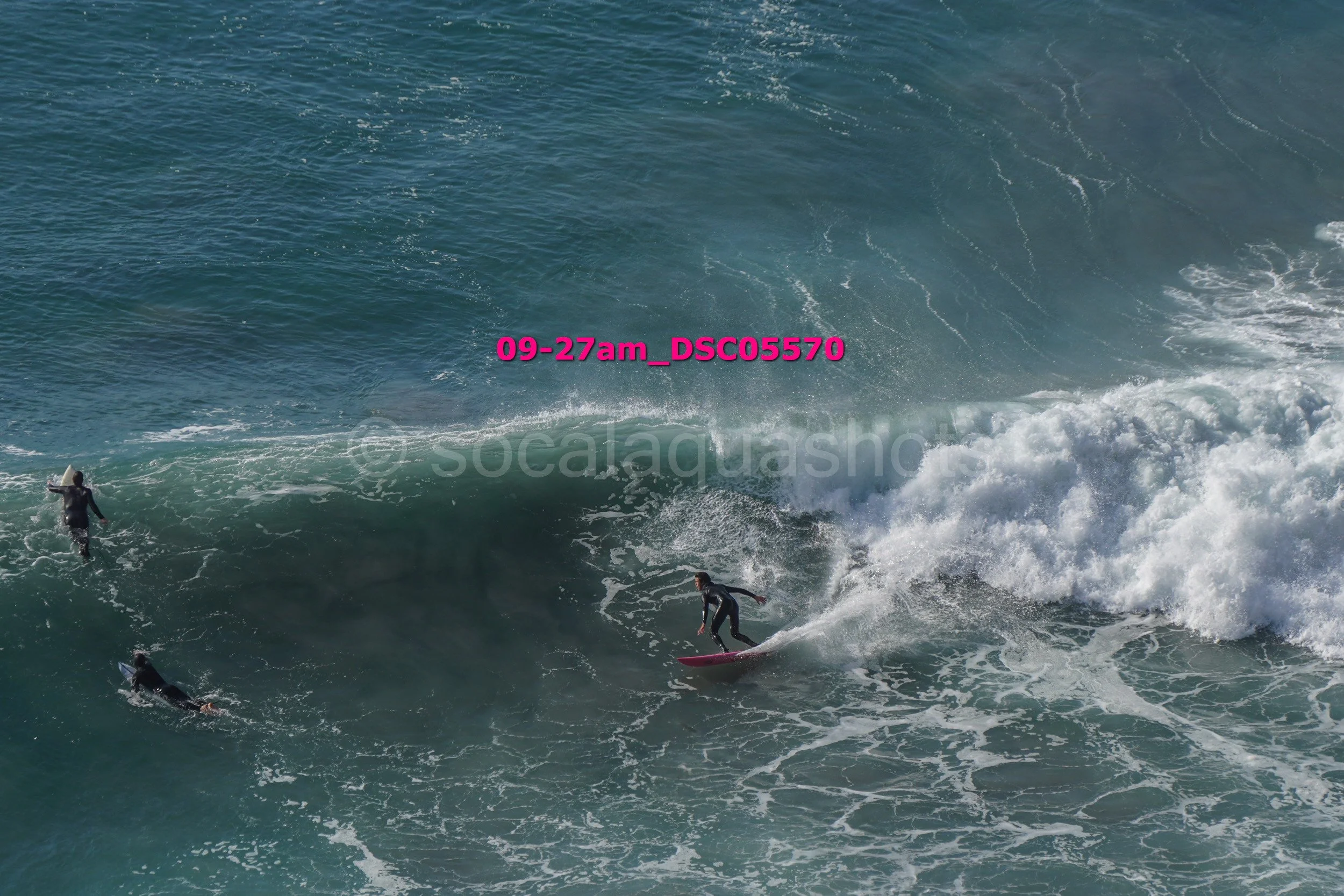 A person surfing on a large ocean wave with two other surfers nearby in the water.
