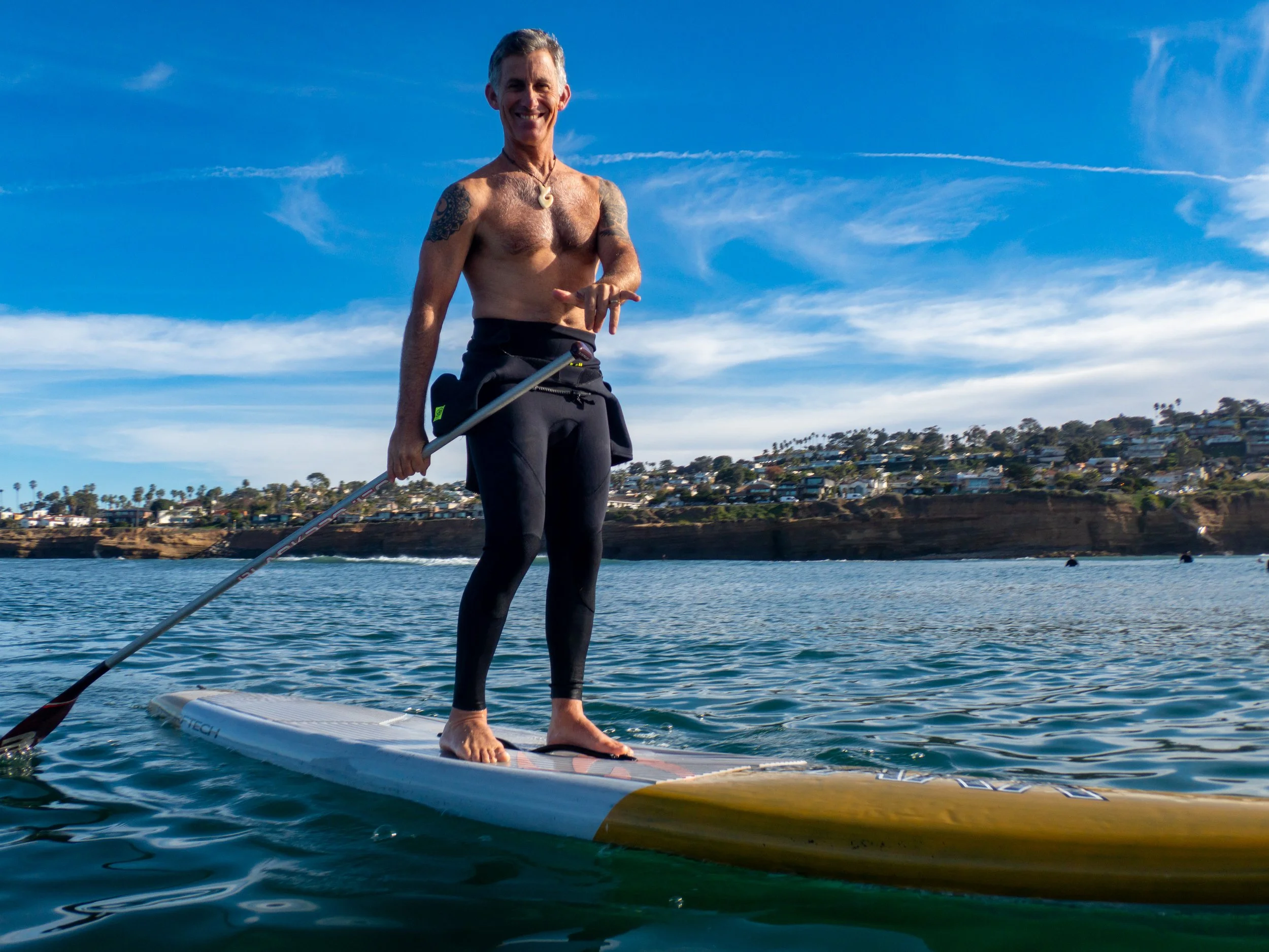 A man paddleboarding on the water under a blue sky with scattered clouds, with a coastline and houses in the background.