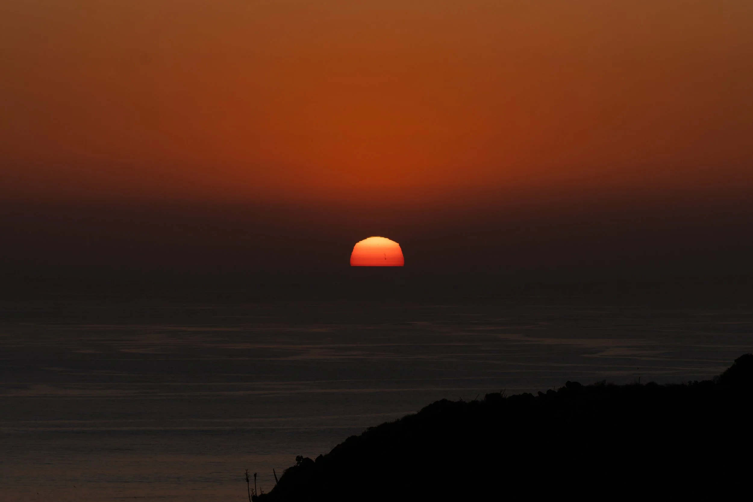 Sunset over the ocean with a dark silhouette of land in the foreground and a vibrant orange sky.