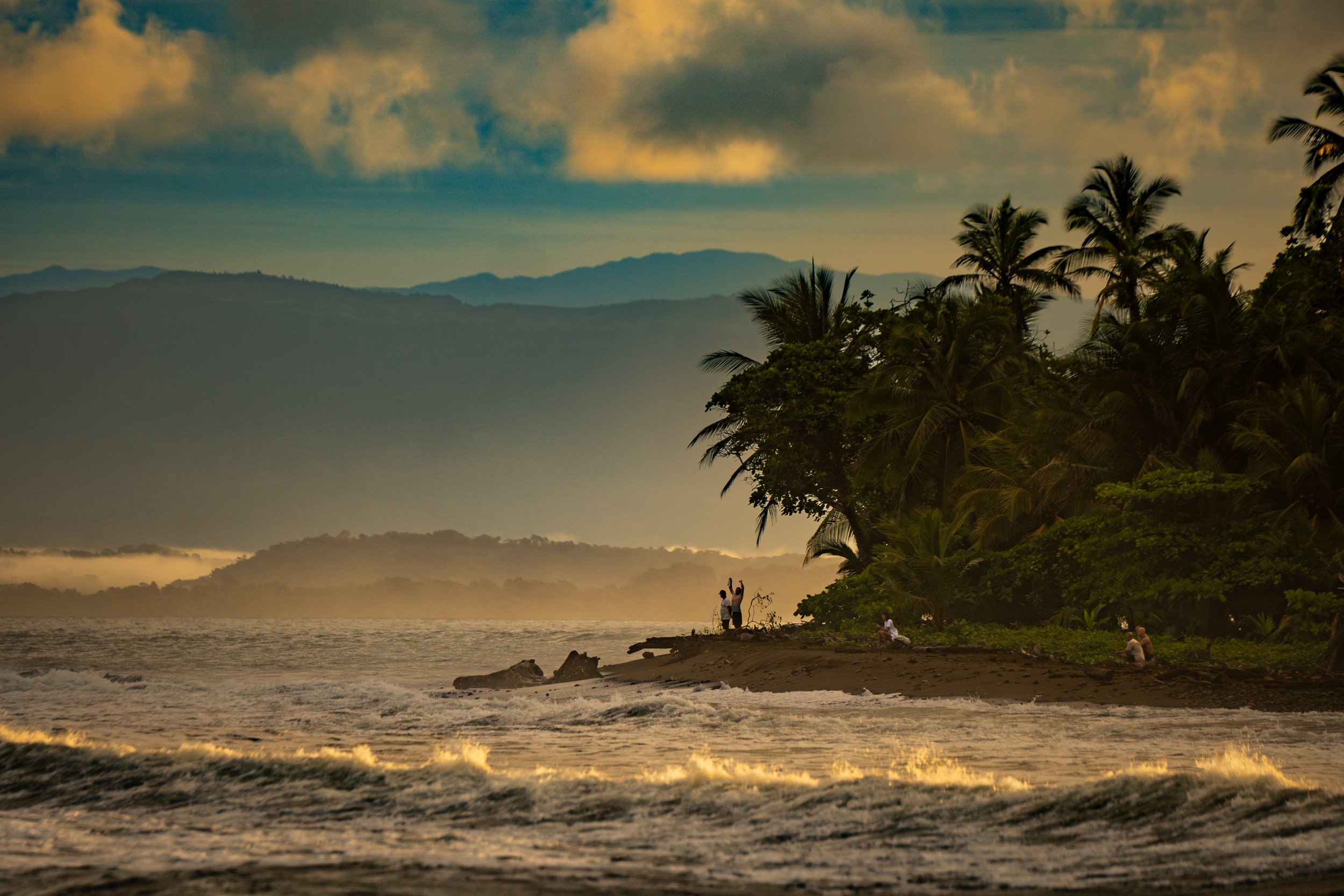 A scenic beach at sunset with palm trees, distant mountains, and a cloudy sky. Several people are enjoying the view near the shoreline, some sitting and some standing.