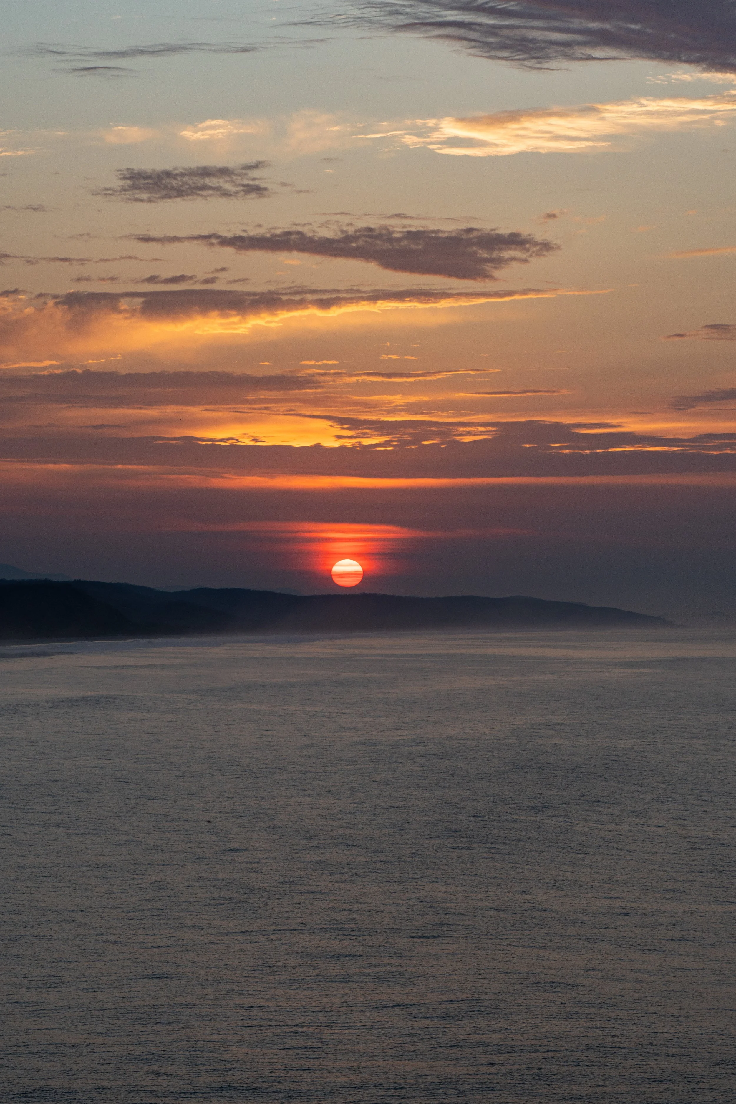 Sunset over a body of water with distant hills, with soft clouds in the sky.