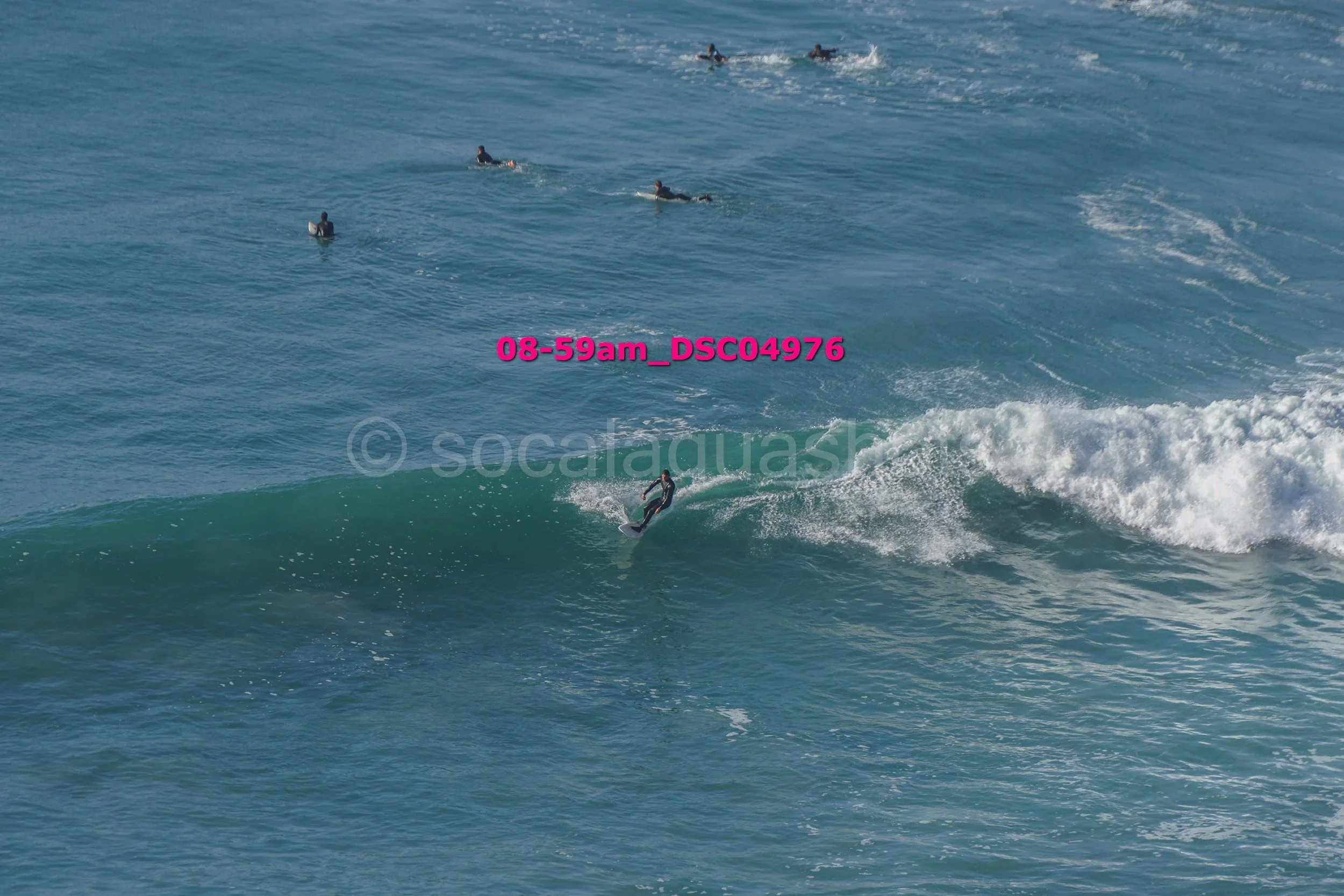 Surfer riding a wave in the ocean with several people swimming in the background.