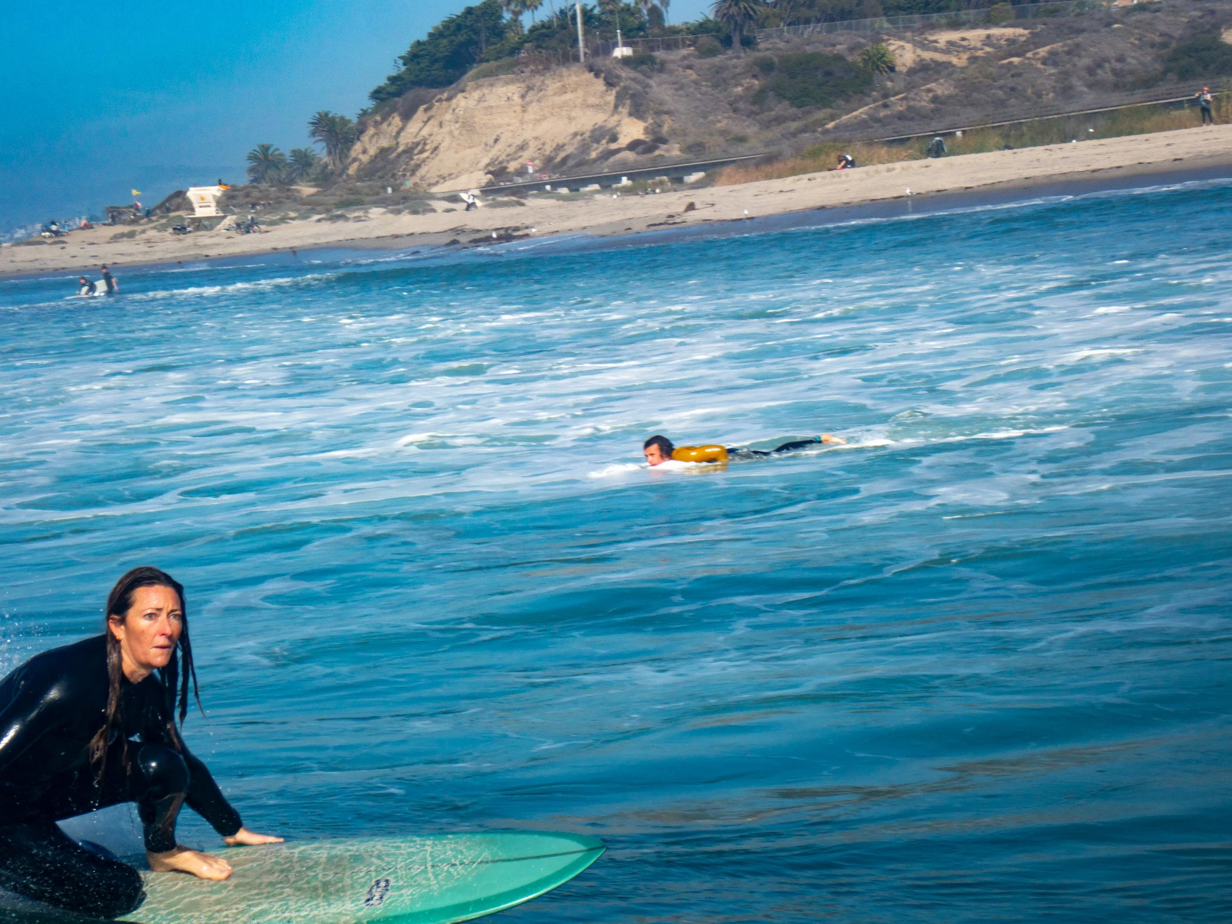 Woman on surfboard in the ocean with another person swimming and a beach in the background.