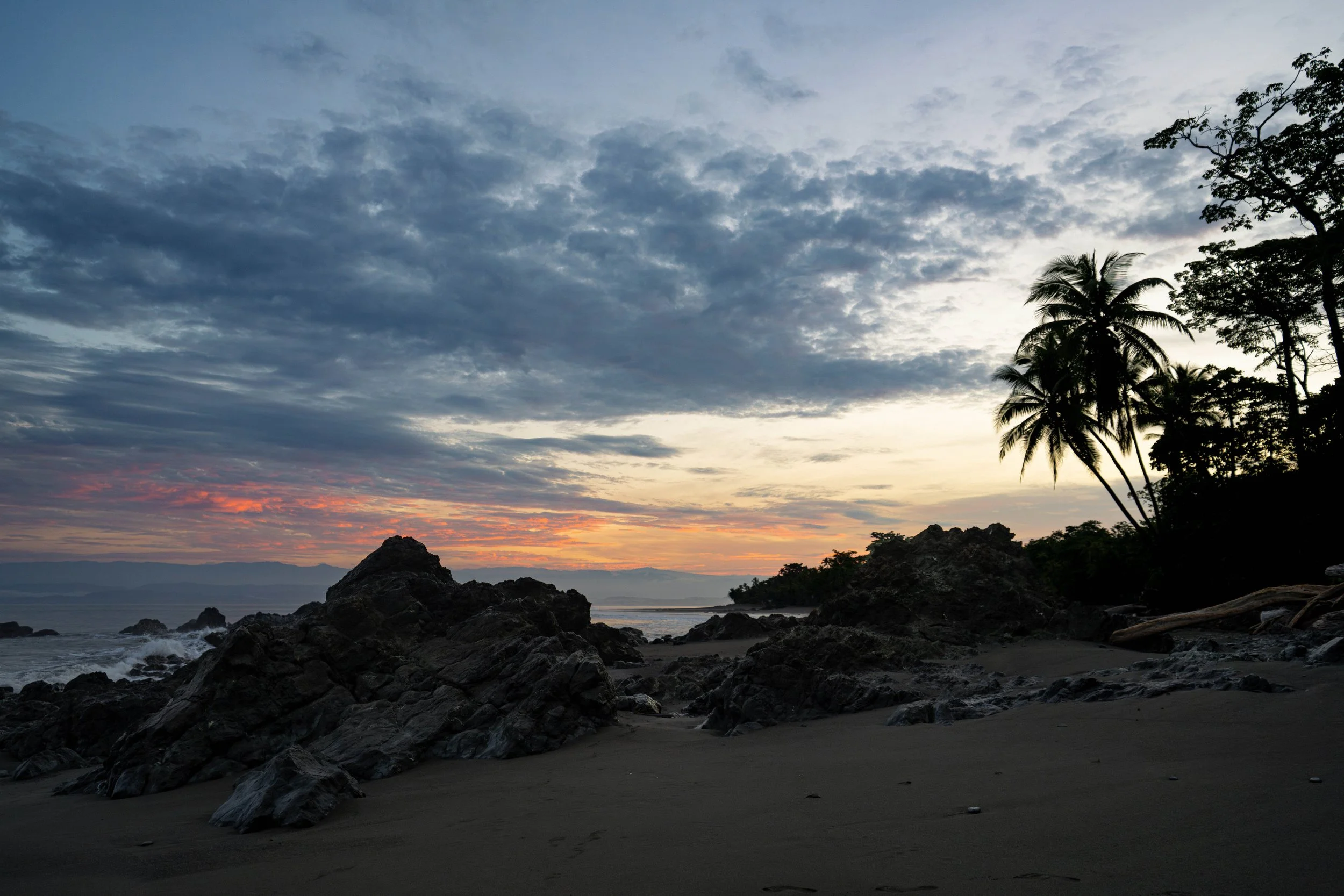 Sunset over a beach with rocks and palm trees, partly cloudy sky, and calm ocean water