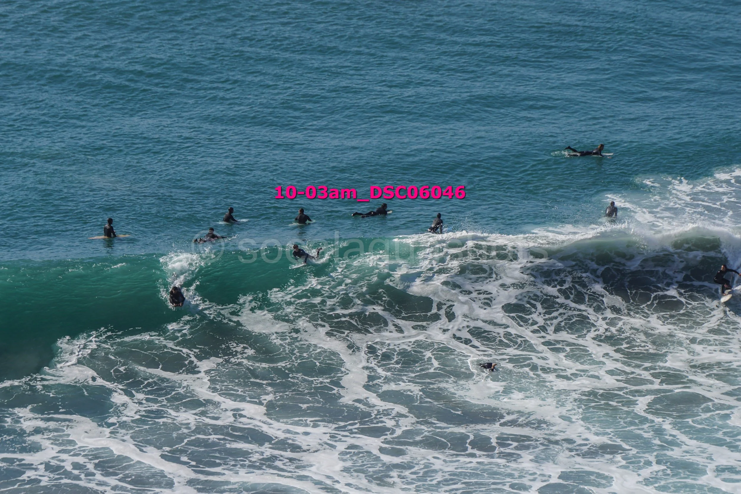 Multiple surfers in wetsuits waiting and riding waves in the ocean, with some in the water and others on surfboards, under clear blue skies.