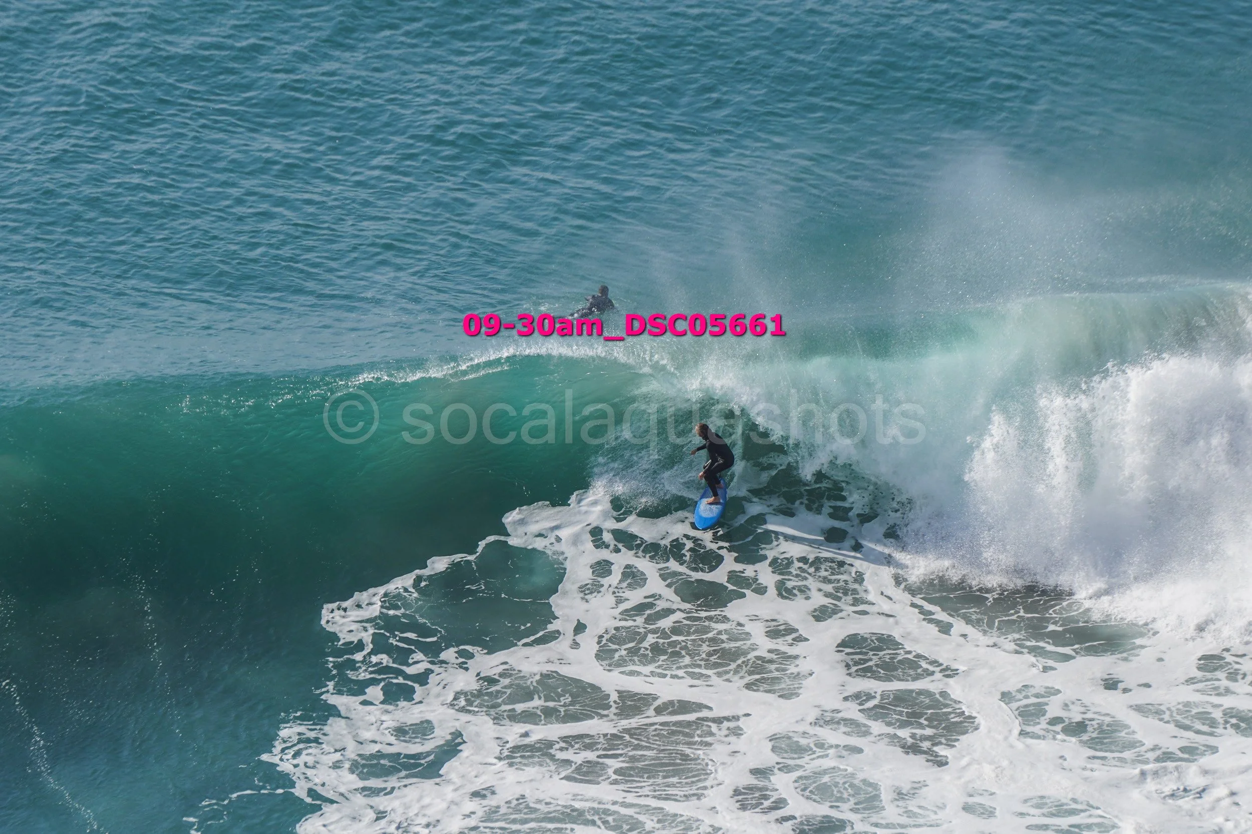 A person surfing on a large ocean wave with another surfer in the background.