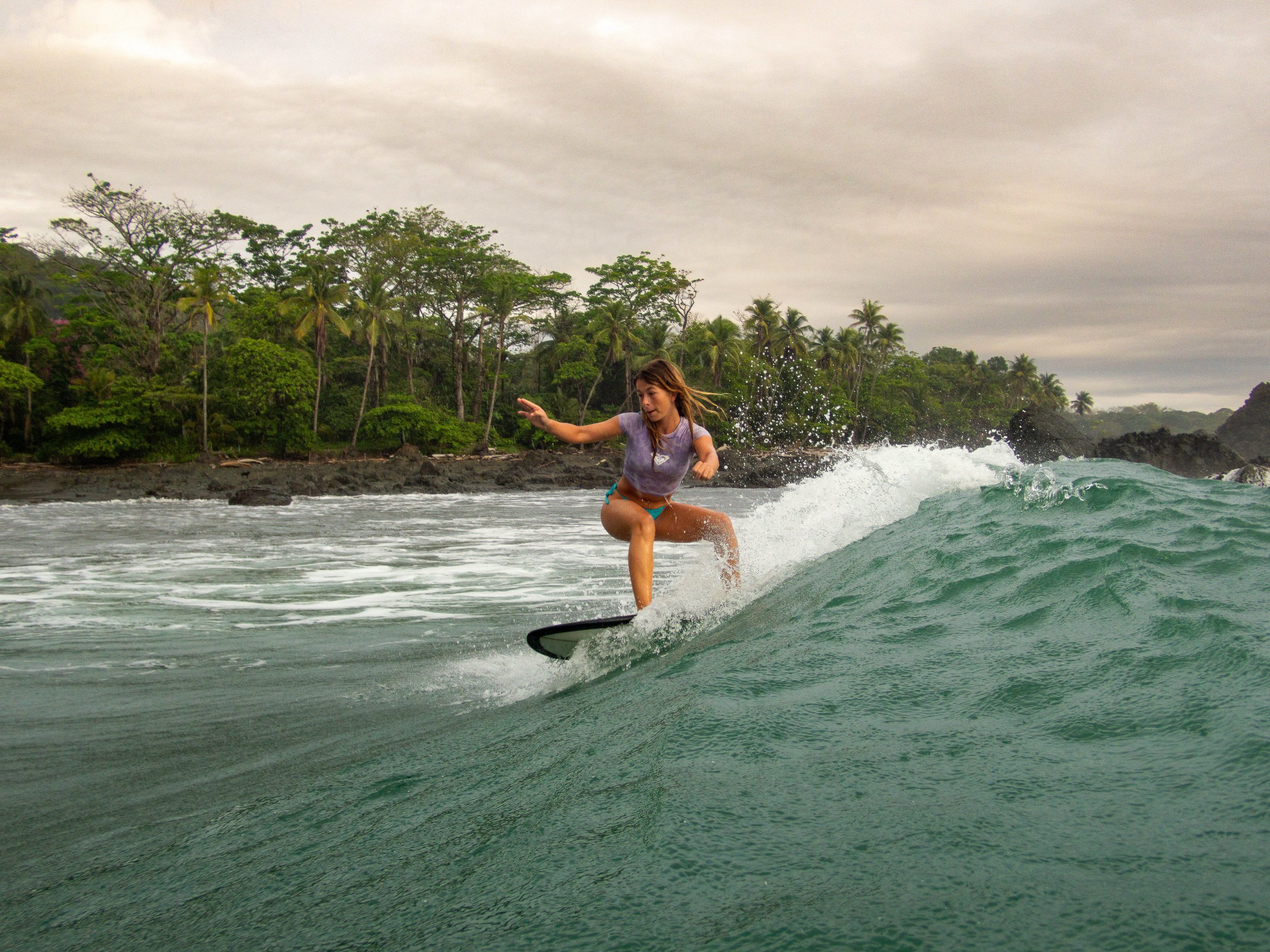 Woman surfing on a wave near a tropical coast with lush green trees.