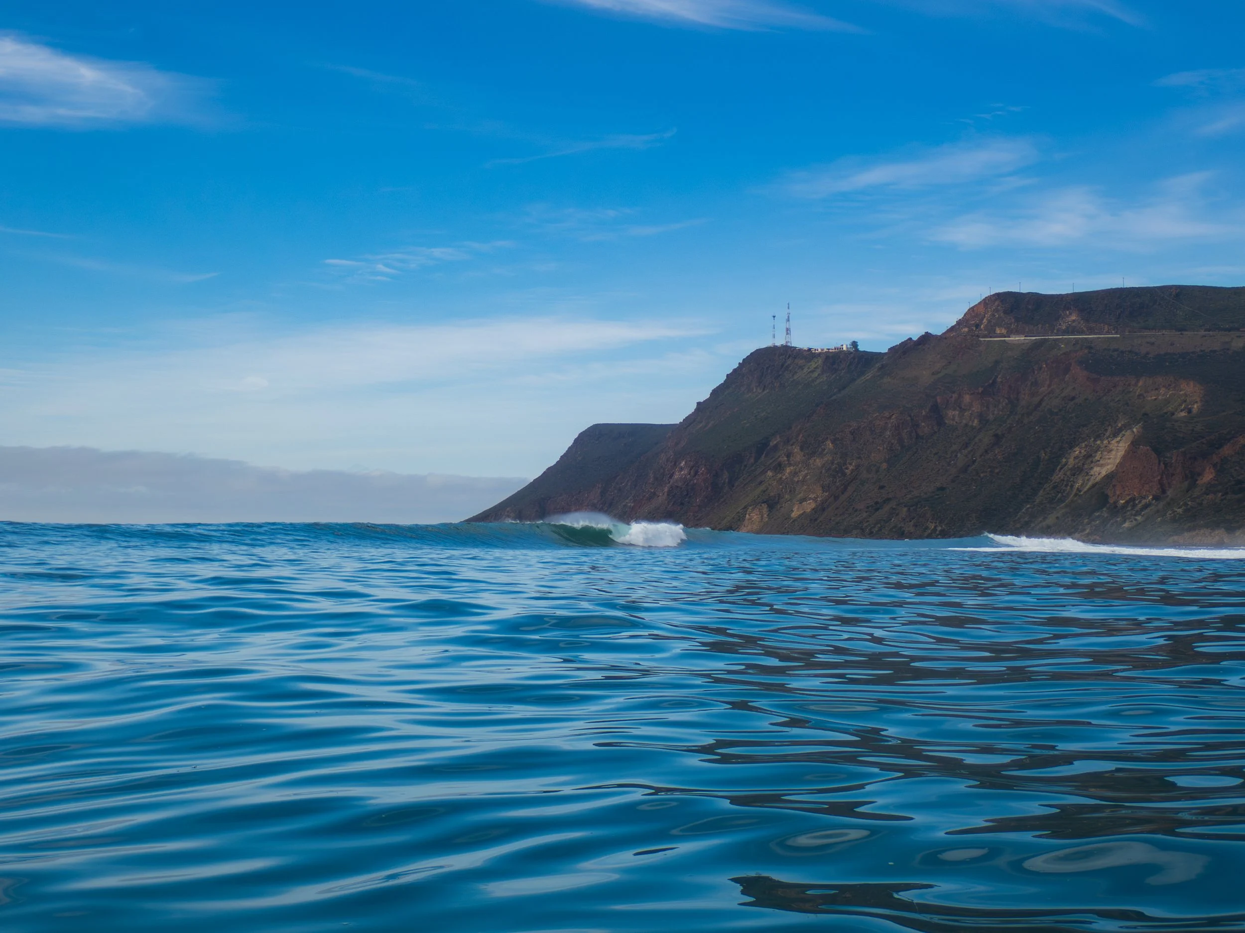 Ocean with small waves near the shoreline, and a mountain or cliff in the background under a blue sky with some clouds.