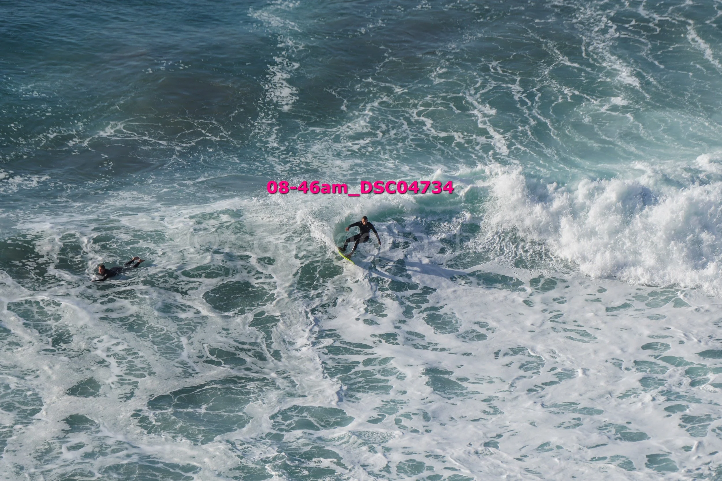 Two surfers riding and lying on ocean waves, with white foam and large water ripples.