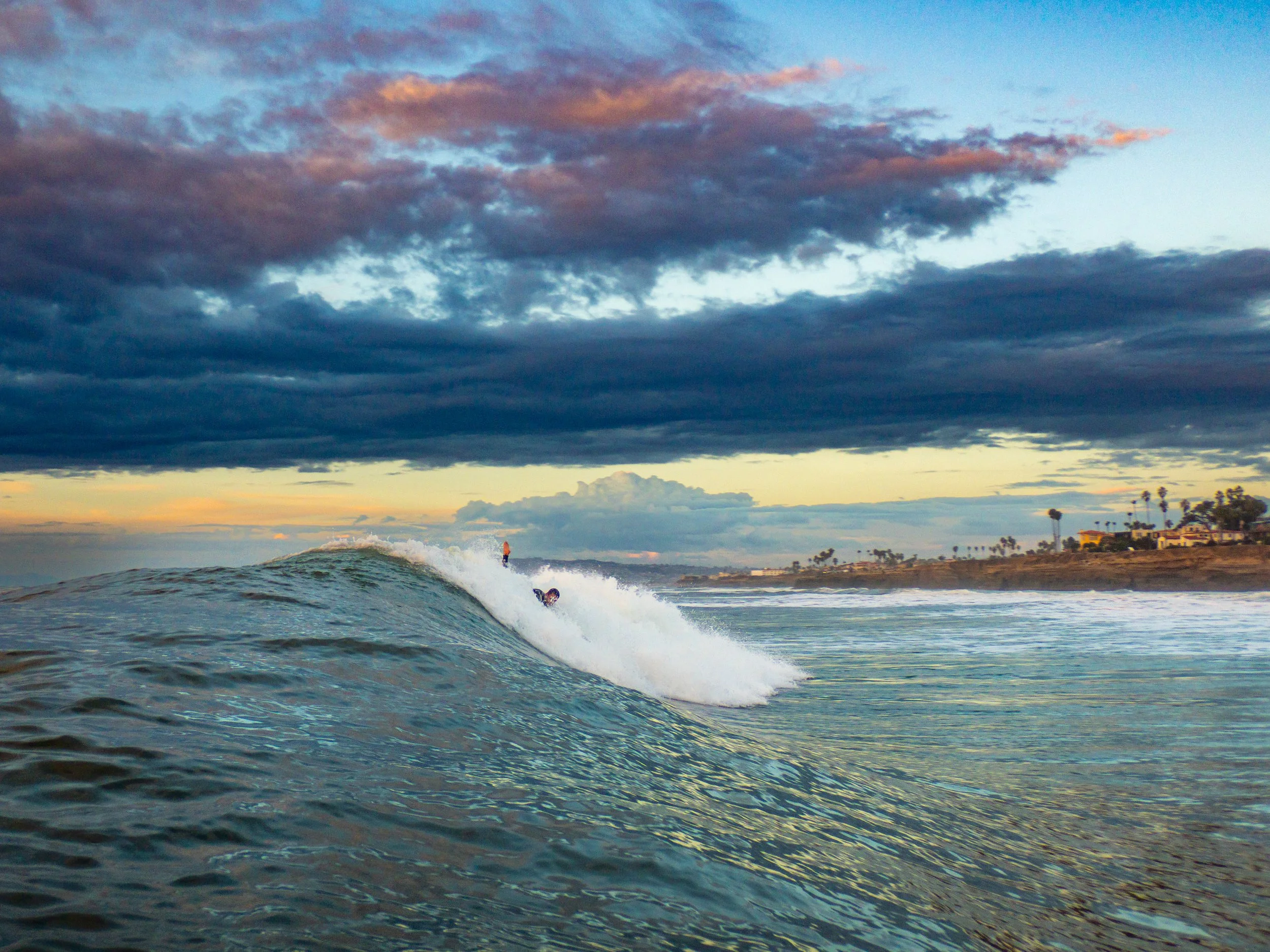 A person surfing on a wave in the ocean during sunset or sunrise with a cloudy sky and a coastal town with palm trees in the background.