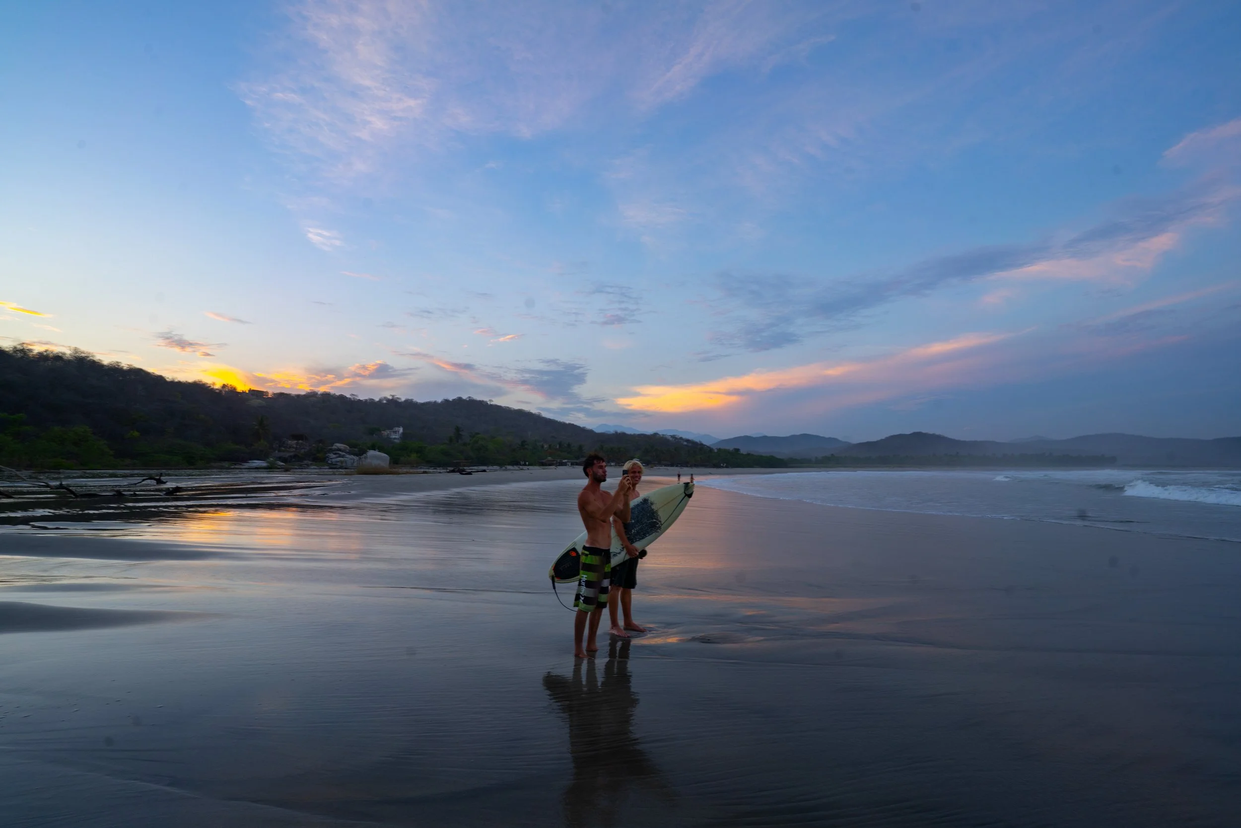 Two men standing on a sandy beach at sunset, one holding a surfboard and the other adjusting his surfboard leash, with mountains and a colorful sky in the background.