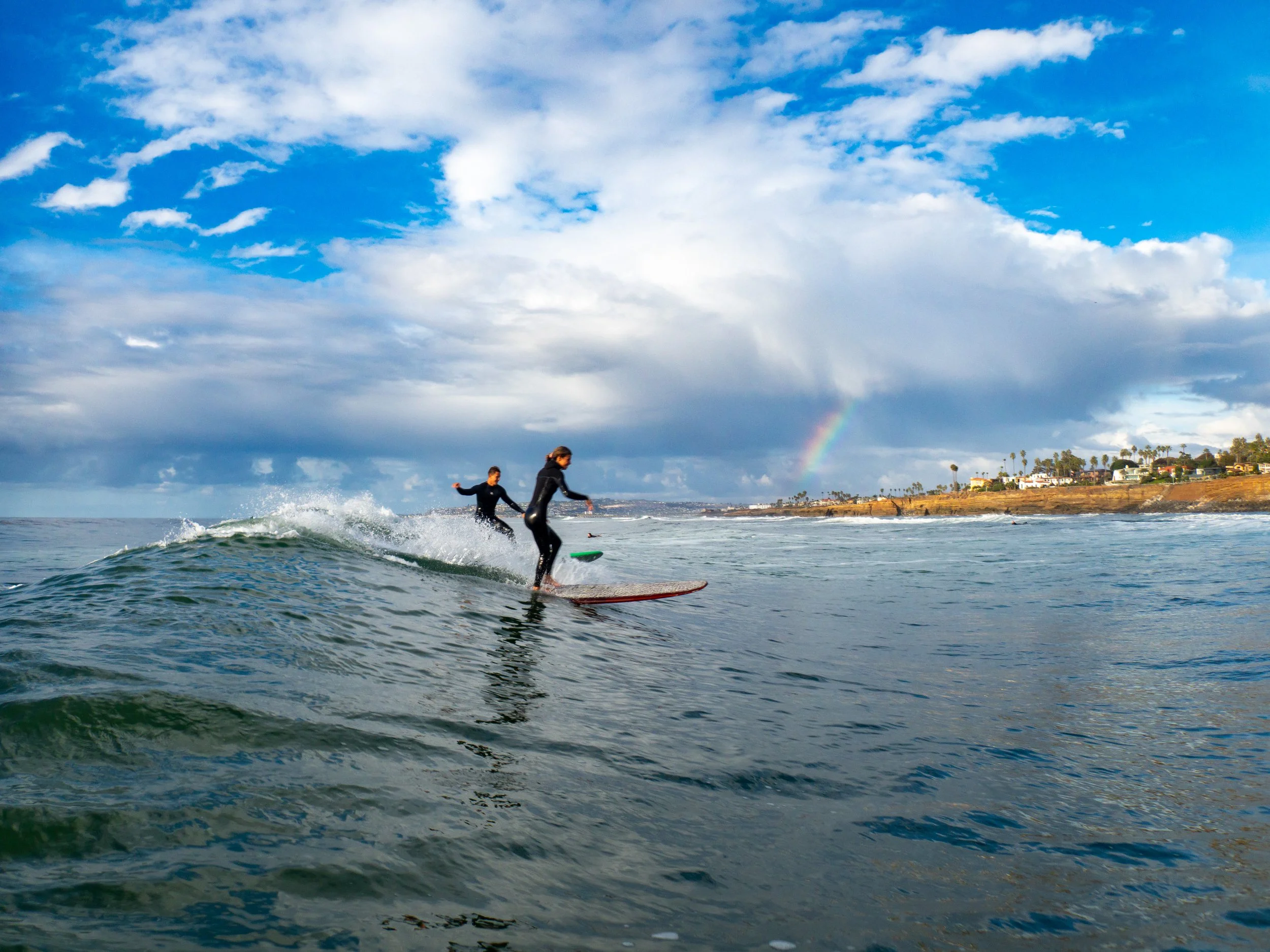 Two people surfing on the ocean with a coastline and palm trees in the background during daytime, with a rainbow in the sky and partly cloudy weather.