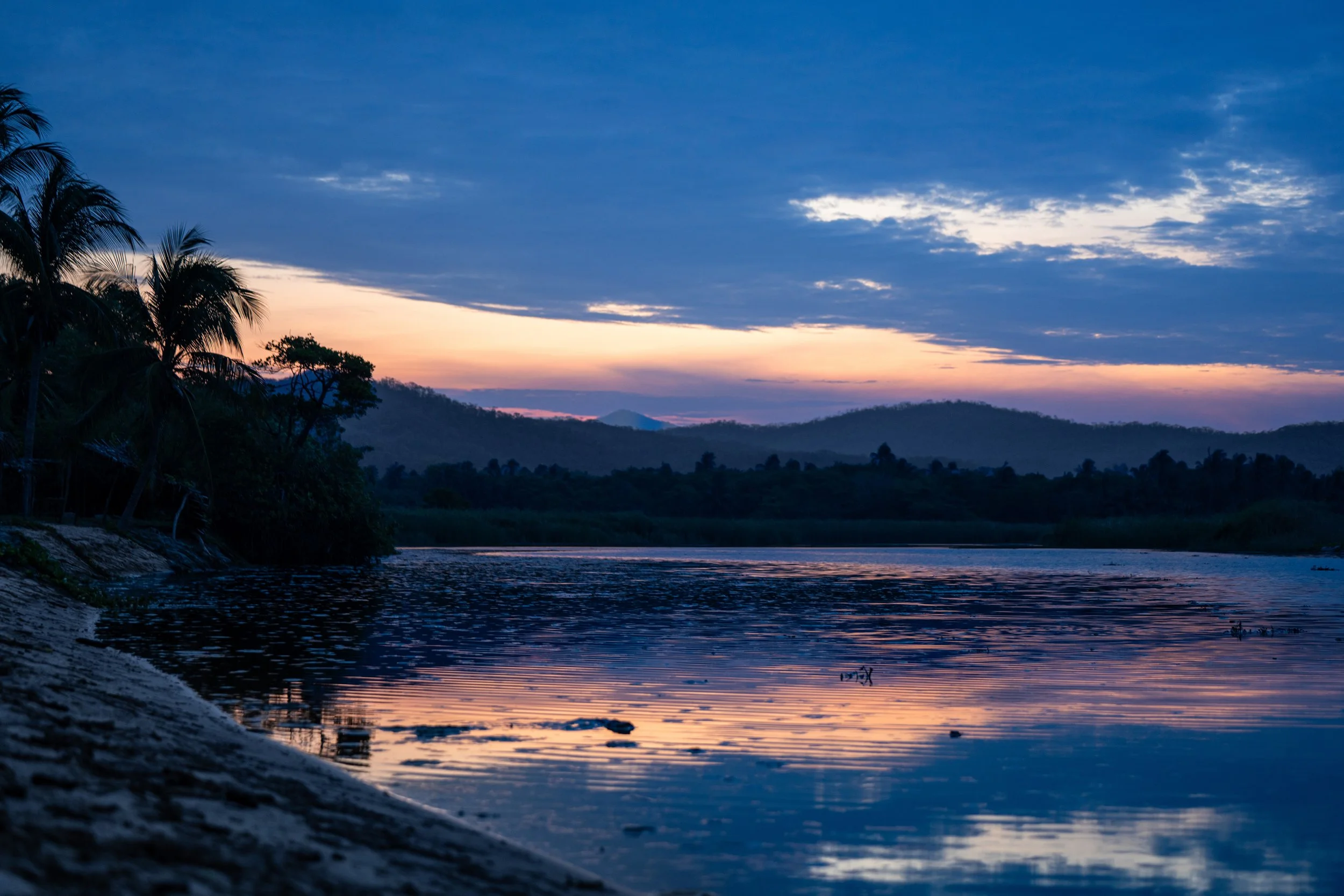 A scenic view of a river at sunset, with dark silhouette trees and hills in the background, and a partly cloudy sky with shades of blue, pink, and orange.