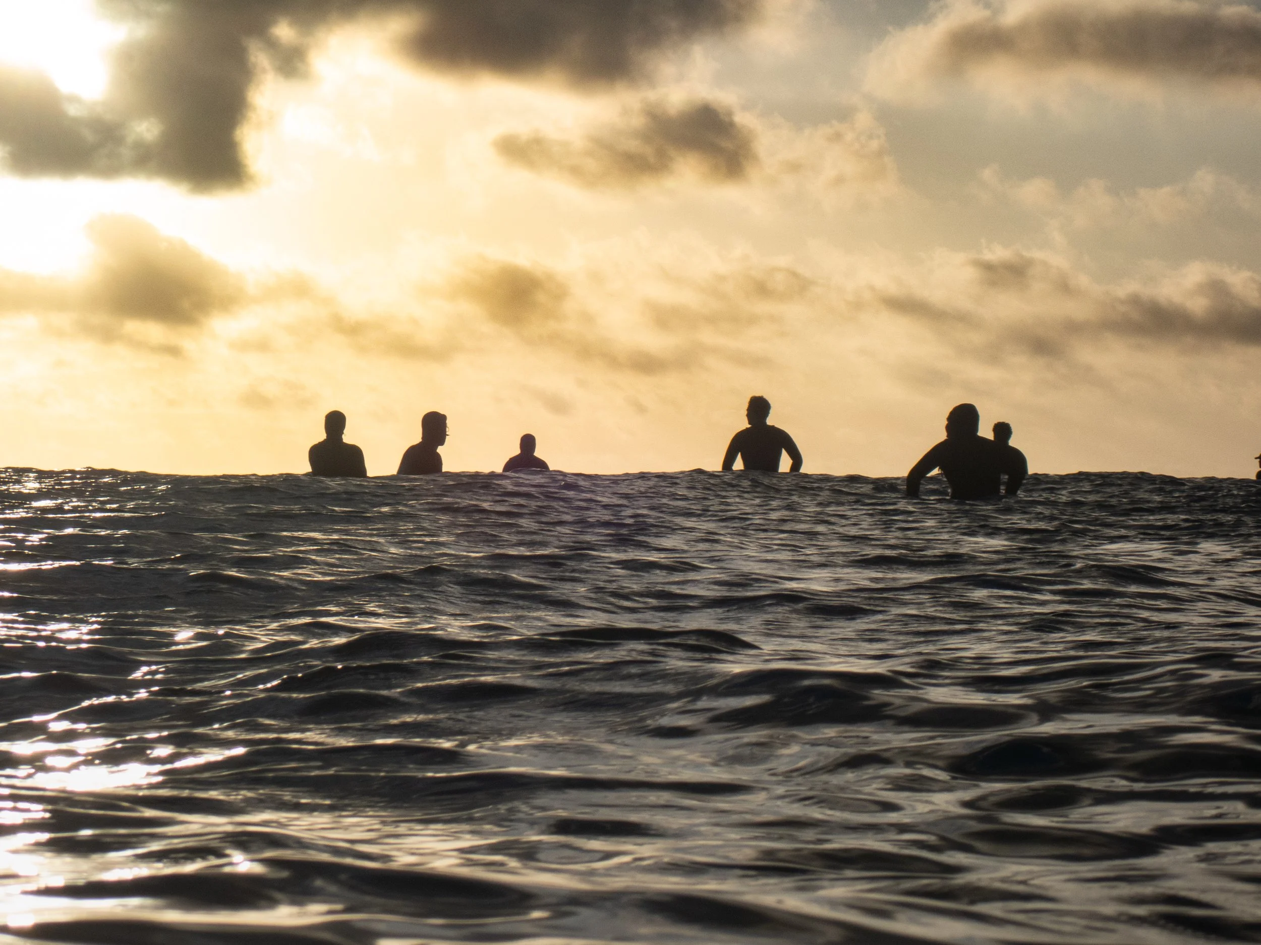 Silhouettes of people in the ocean at sunset with clouds in the sky.