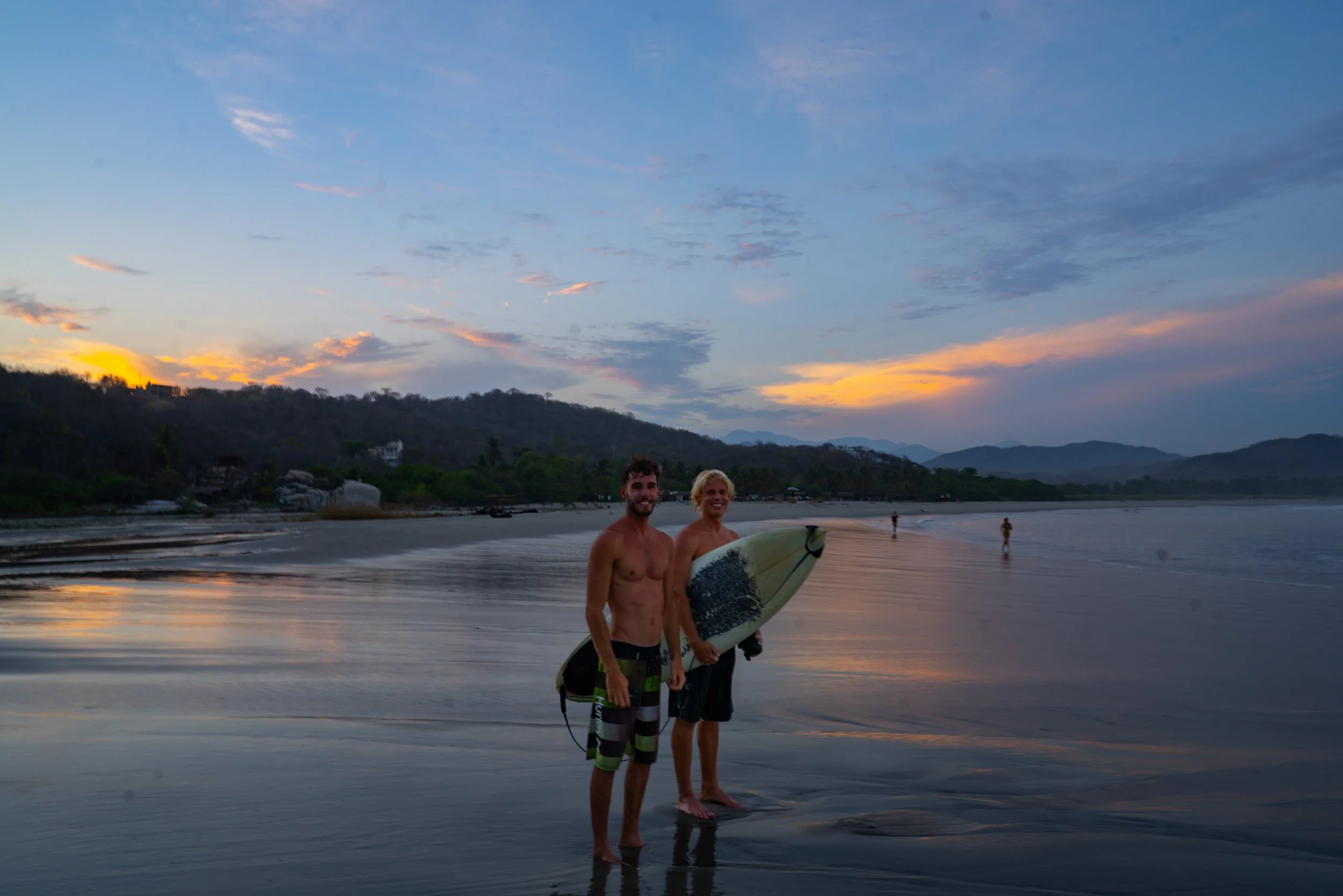 Two shirtless men holding surfboards standing in shallow water at sunset on the beach, with a scenic background of distant hills and a sky with scattered clouds.