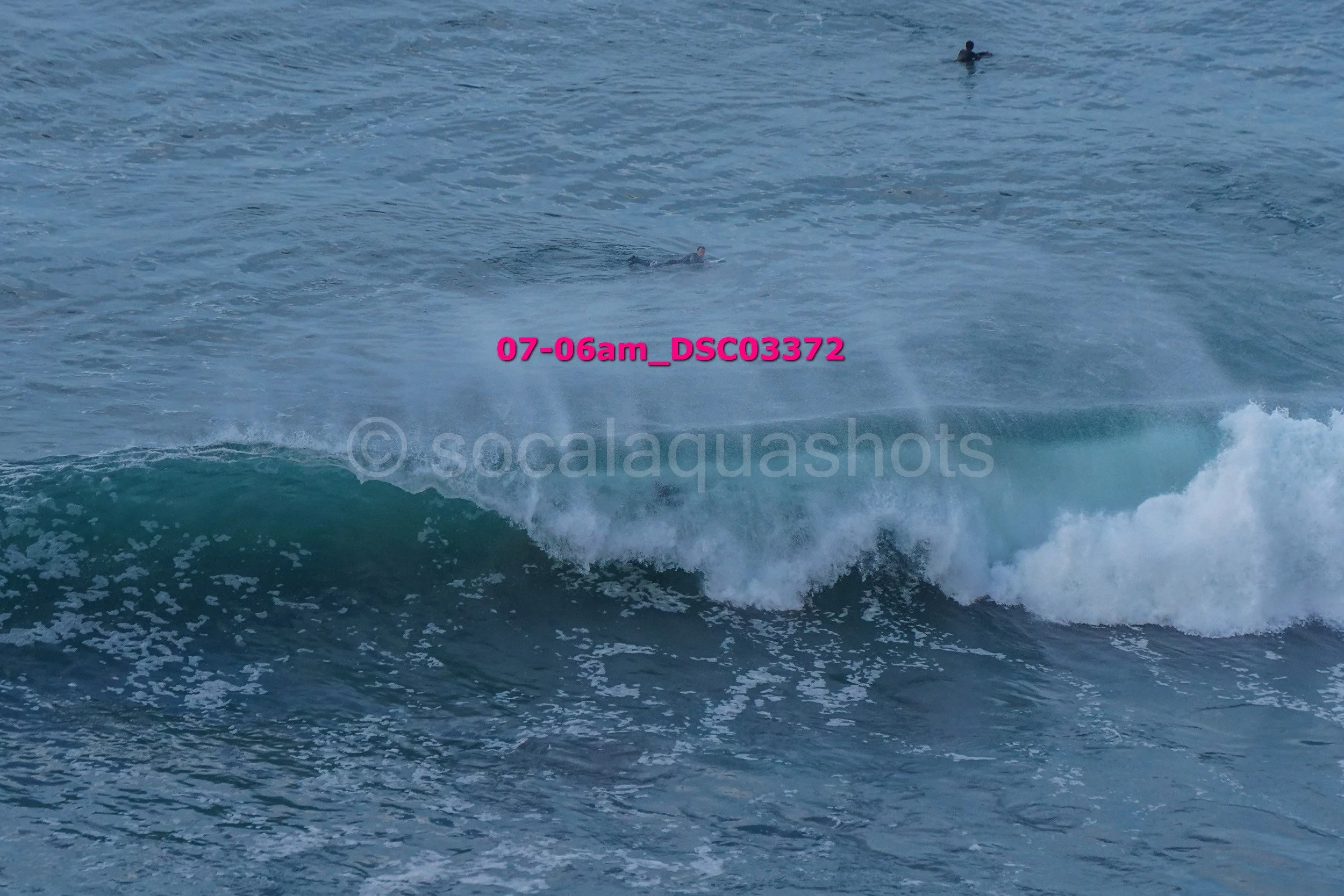 Ocean waves with surfers in the distance, one visible on a surfboard riding a wave.