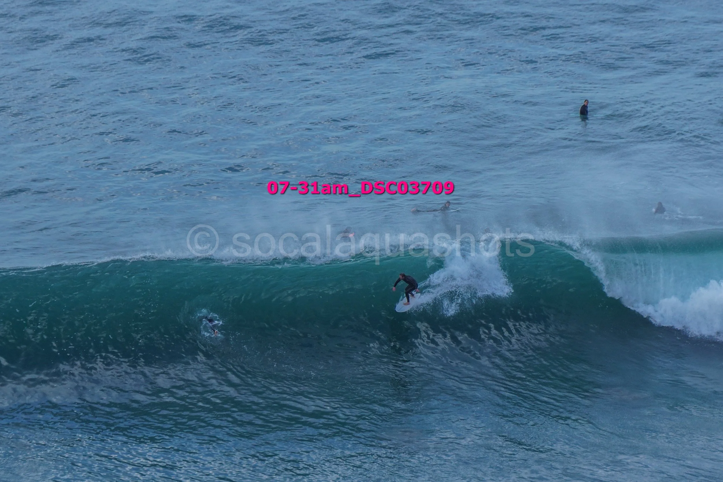 Surfer riding a wave in the ocean with other surfers around, some in the water and others standing or floating up ahead.