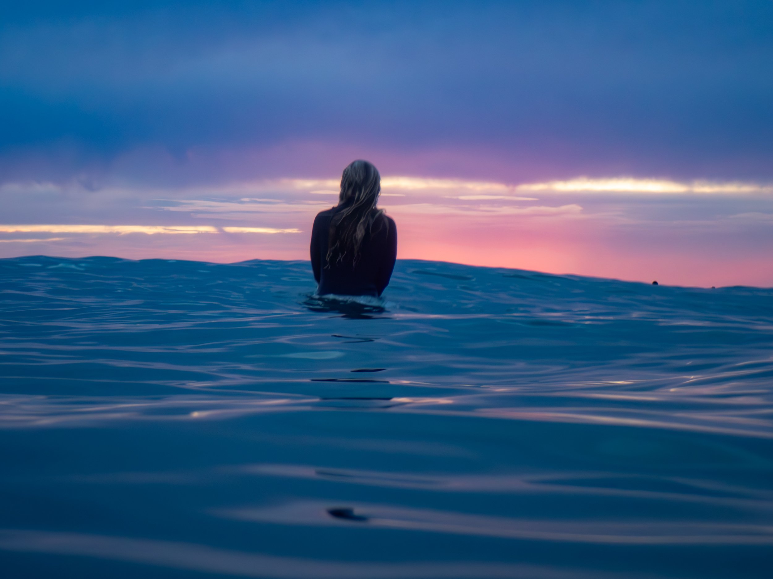 A woman standing in the ocean at sunset, facing the horizon with purple and pink skies.