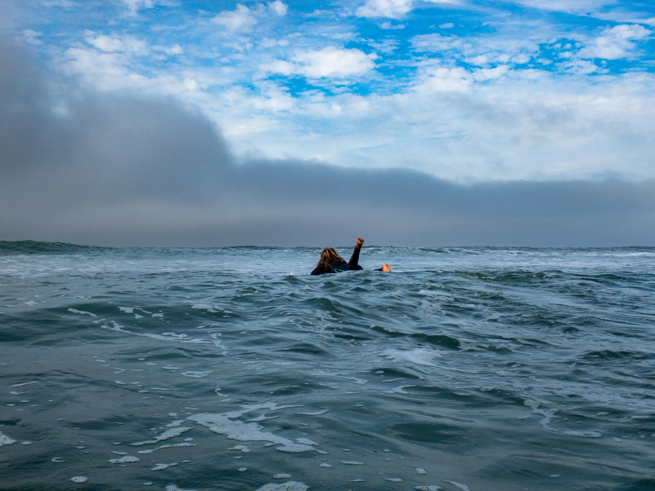 A person in the ocean, possibly swimming or snorkeling, with their arm raised. The sky is partly cloudy with blue skies and some dark clouds in the distance.